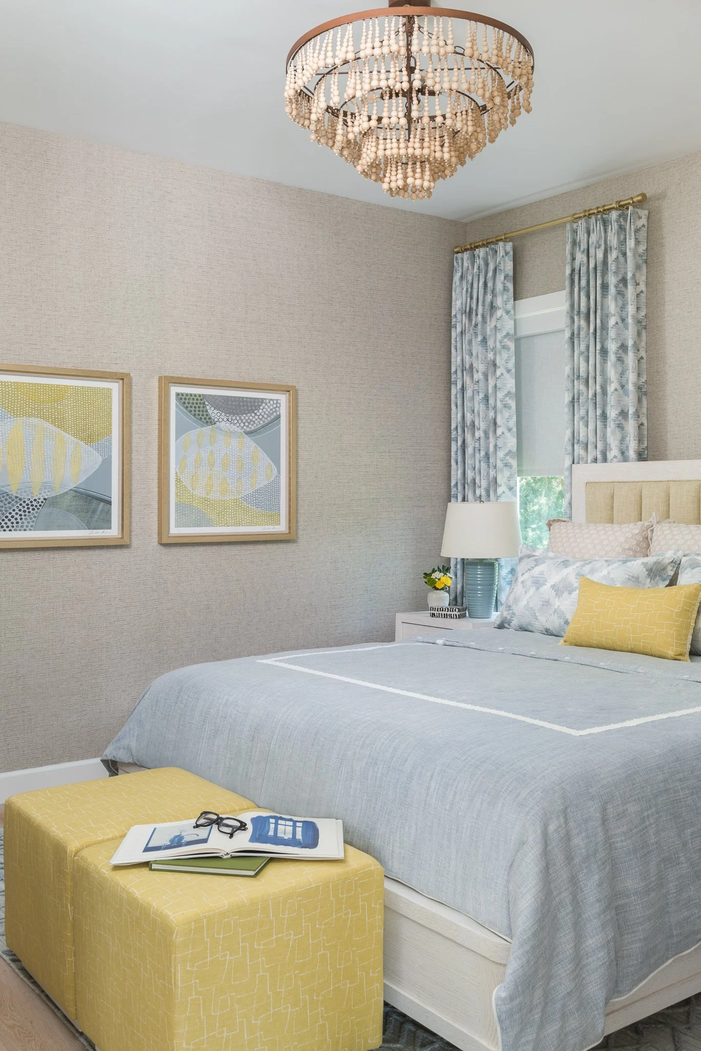 A bedroom with a bed, yellow ottoman, and windows with patterned curtains, adorned with artwork and a chandelier.