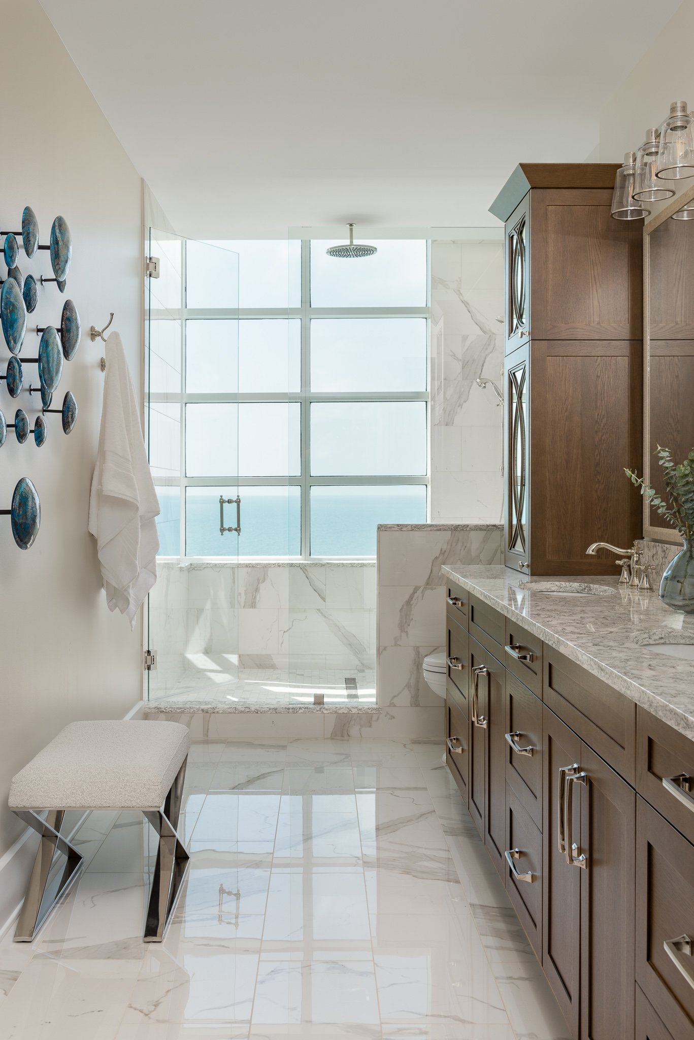 A luxurious bathroom with marble floors and walls, a large walk-in shower with glass doors, wooden cabinetry, and a window with a view of the sky and ocean.