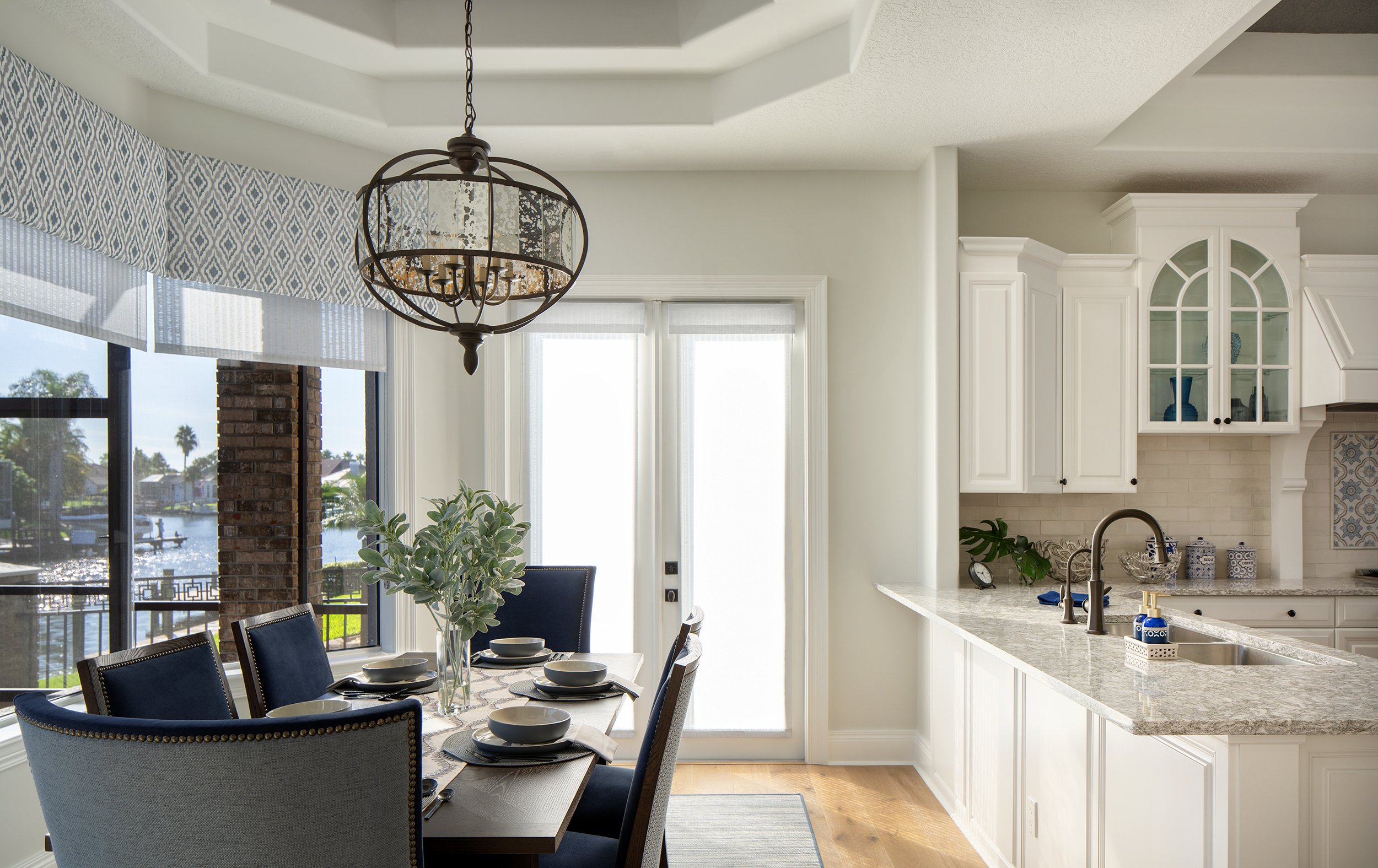 Bright dining area with a table set for six, black chairs, a large window showing a waterfront view, and a white kitchen with granite countertops and glass-front cabinets.