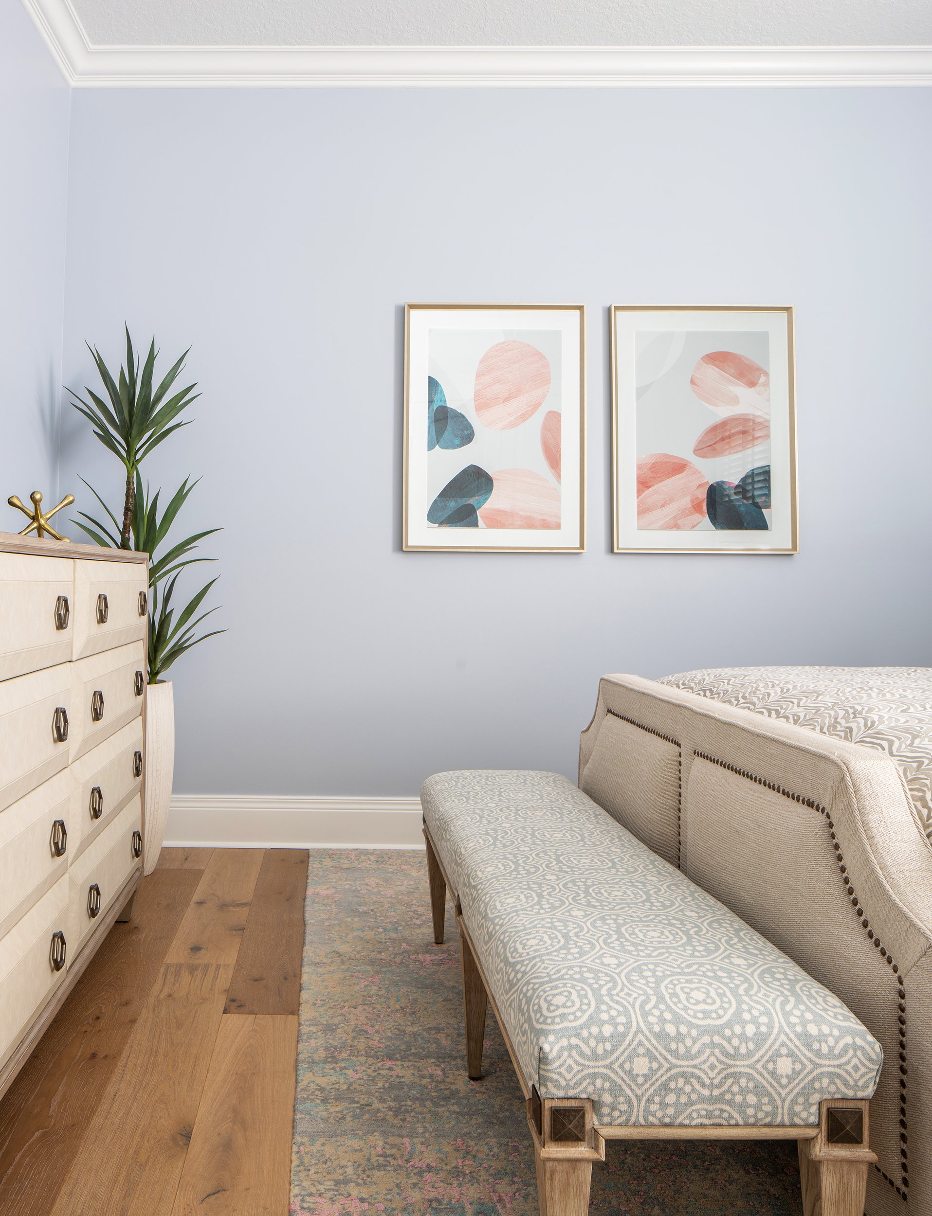 A bedroom with a beige upholstered bed, a patterned bench at the foot, a wooden dresser with brass handles, a large green plant, and two abstract framed art prints on a light blue wall.