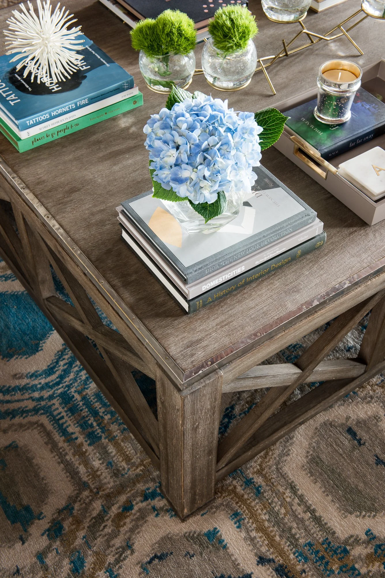 Rustic wooden coffee table with books, a blue hydrangea in a glass vase, two small plants in round glass containers, a lit candle, and a tray of objects. Part of a blue patterned rug is visible underneath.