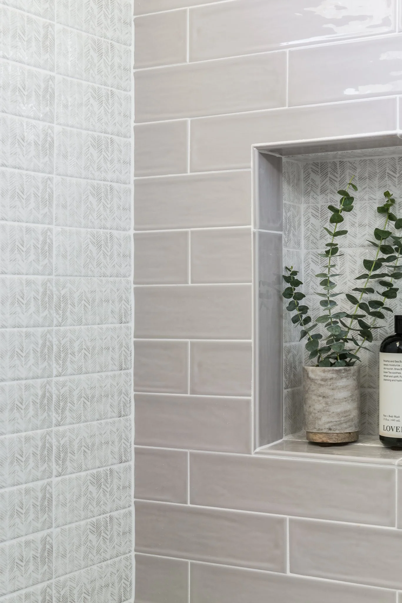 Close-up of a tiled bathroom wall with a built-in shelf holding a potted plant and a bottle of soap.