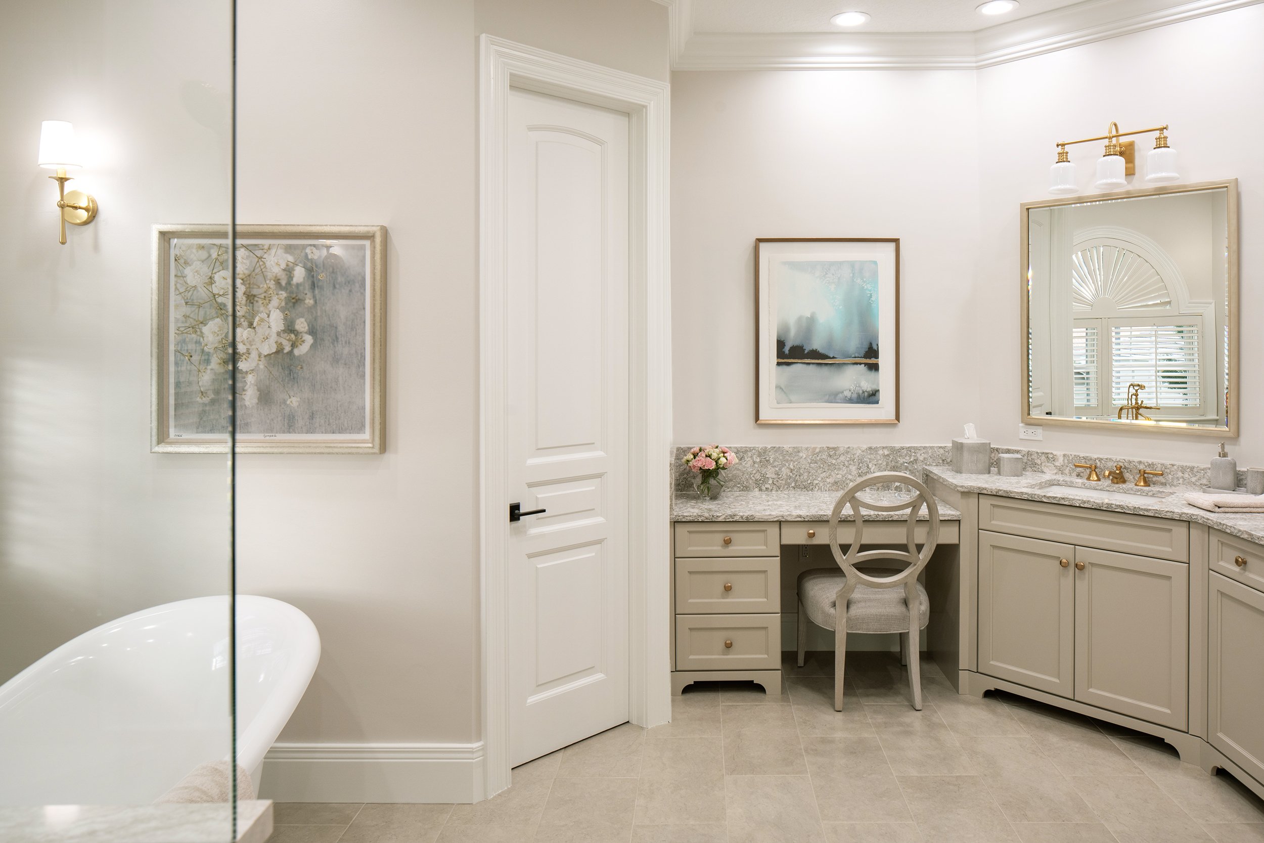 Bright, elegant bathroom with cream-colored cabinetry, granite countertops, a large mirror, framed artwork, a small vanity with a chair, and a bathtub partially visible in the foreground.