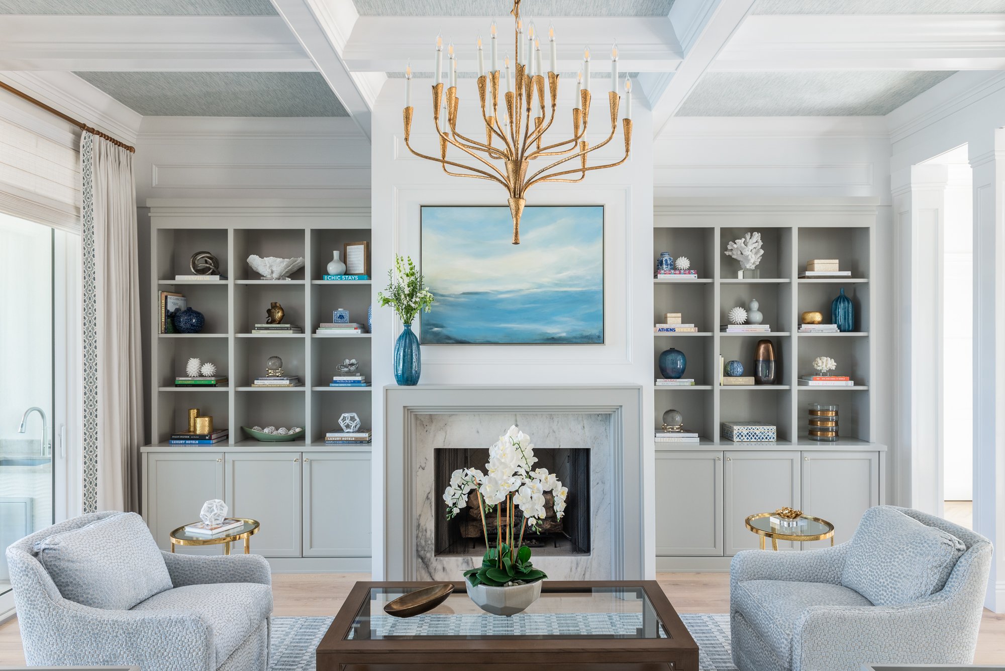 A living room with white walls, built-in bookshelves filled with decorative items and books, a marble fireplace with a potted orchid on the mantel, and a large abstract seascape painting above it. The room features two light gray armchairs, a wooden 