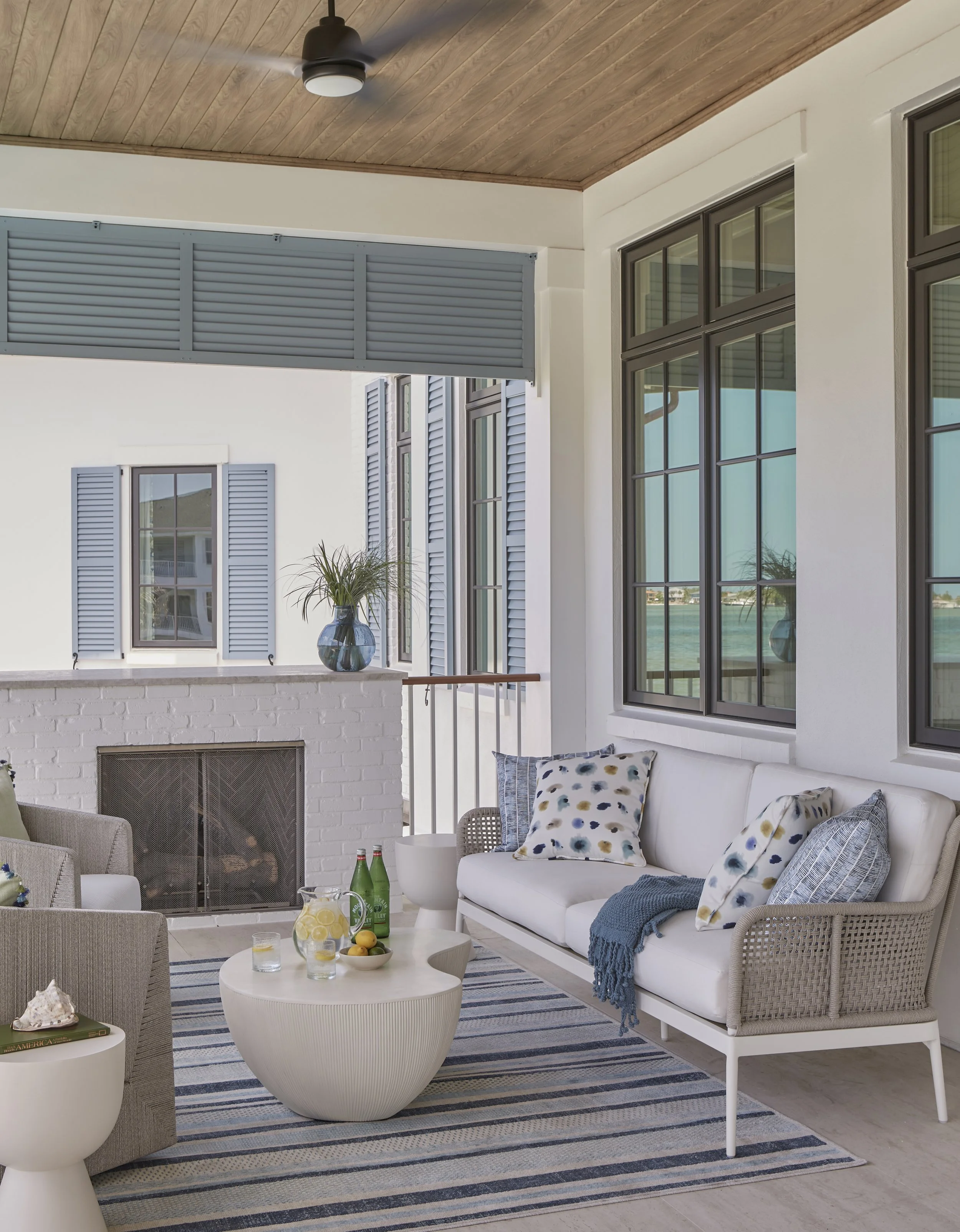 Outdoor porch with white cushioned seating, blue and white patterned pillows, a striped rug, a white round coffee table with lemonade and bottles, a fireplace with a blue vase and greenery, and a view of water through large windows.