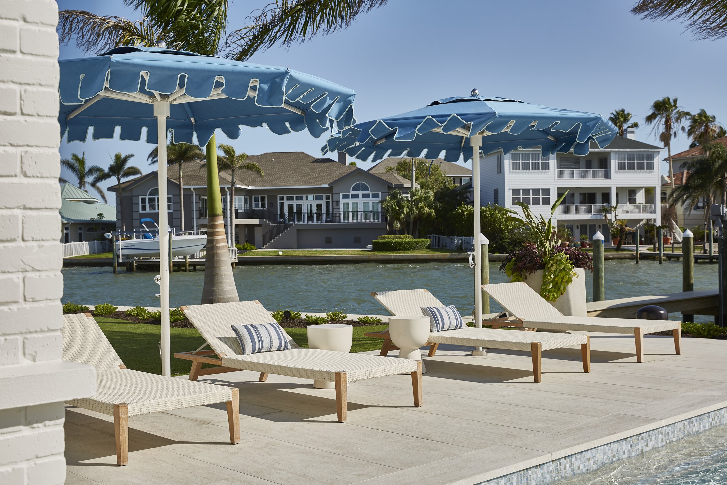 Poolside area with lounge chairs, umbrellas, and waterfront houses in the background under a clear blue sky.