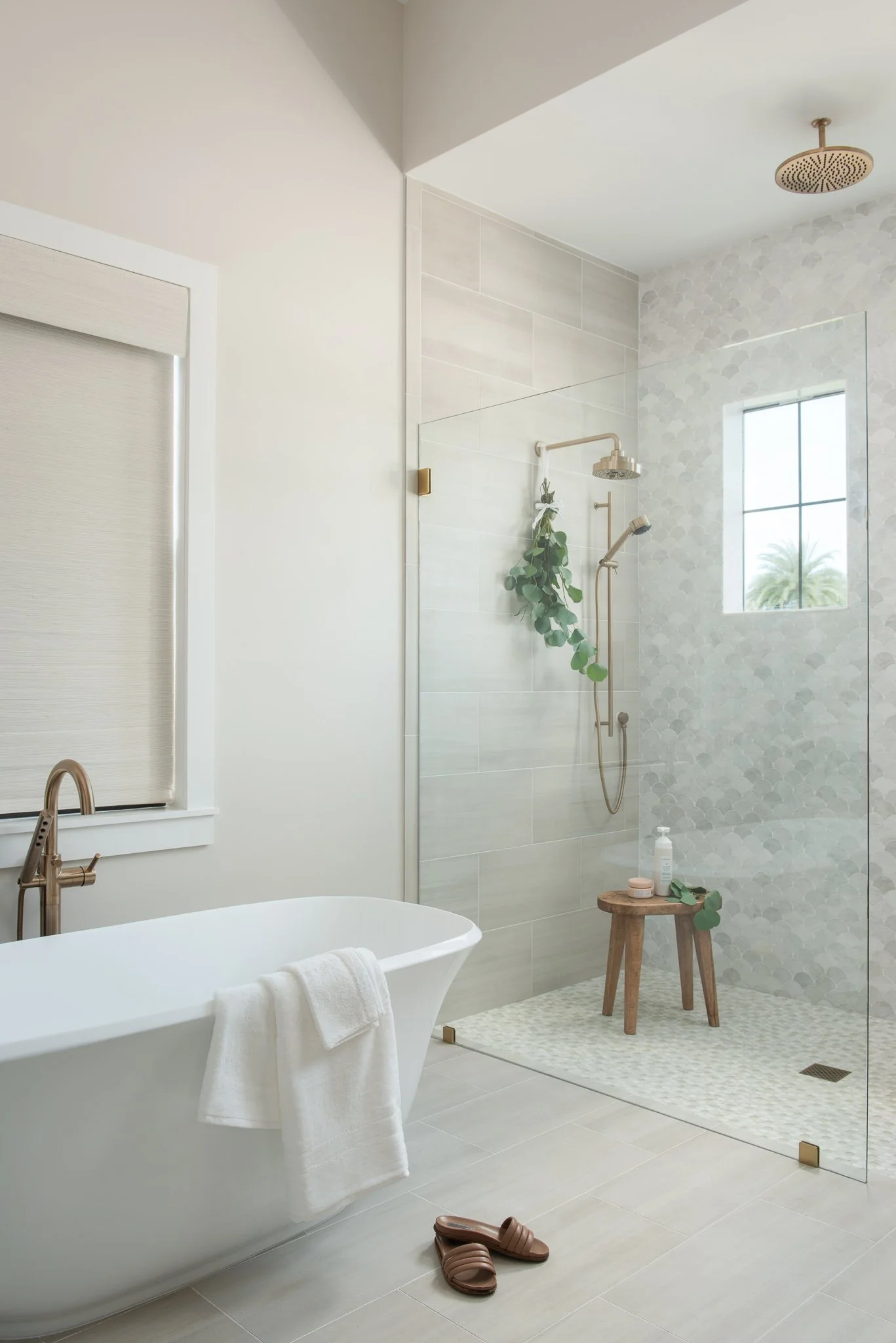 Modern bathroom with a white freestanding bathtub, brown slippers on the floor, and a walk-in shower with a wooden stool, green plant, and a window showing palm trees outside.