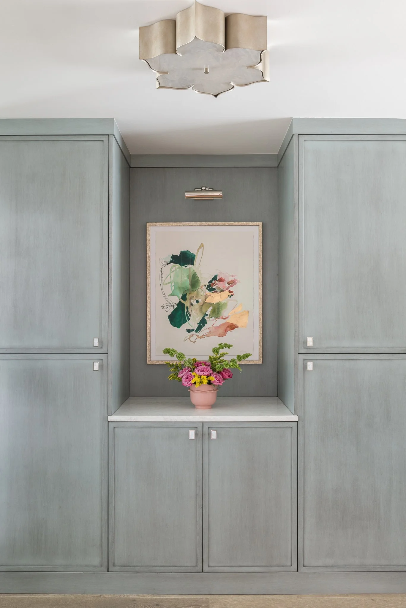 A built-in light grey cabinet with a framed abstract watercolor art on it, a pink vase with pink and yellow flowers, and a modern ceiling light fixture in a minimalistic interior.