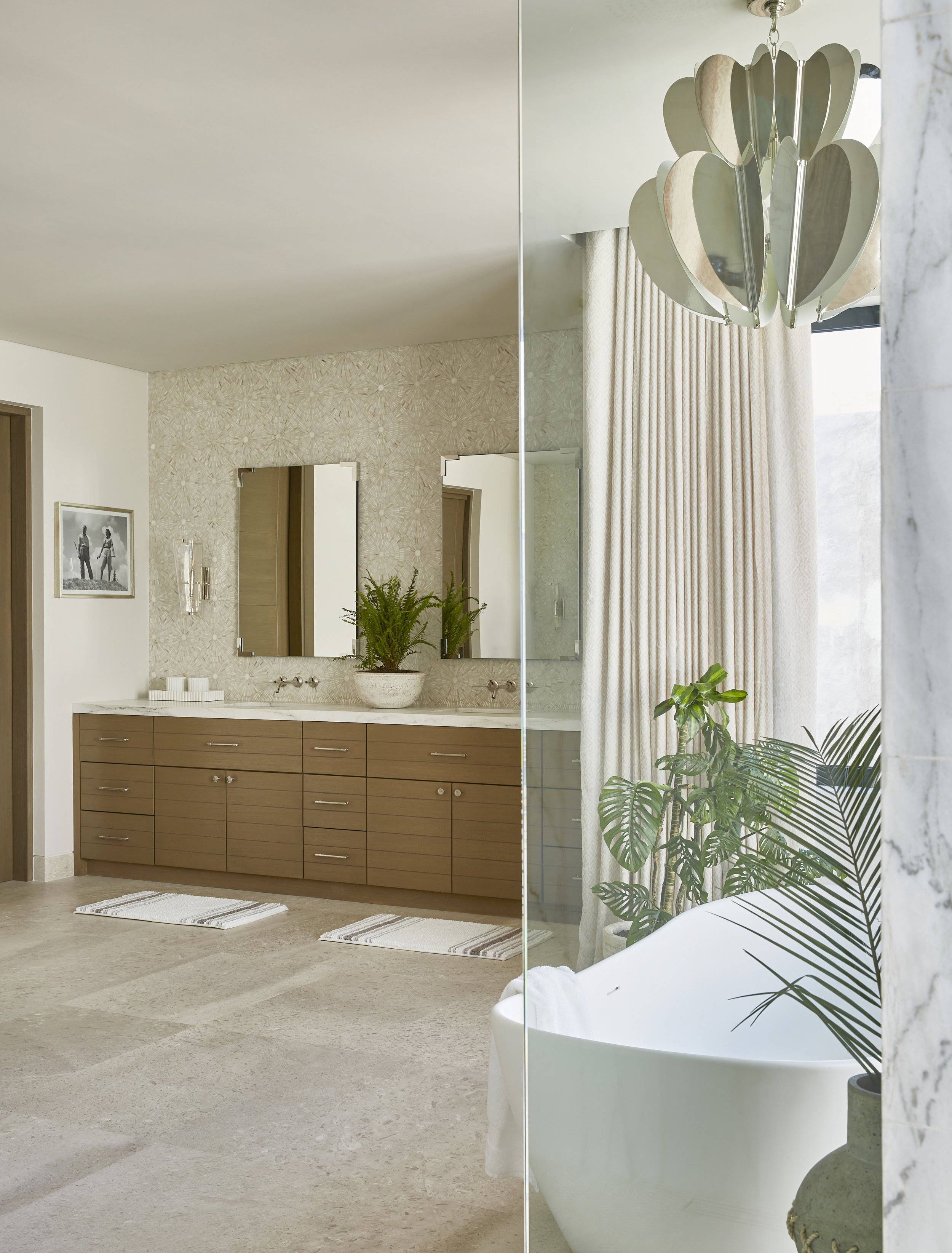 Modern bathroom with a large mirror, double sinks, wooden cabinets, decorative wallpaper, a bathtub with a white towel, indoor plants, beige curtains, and a chandelier.