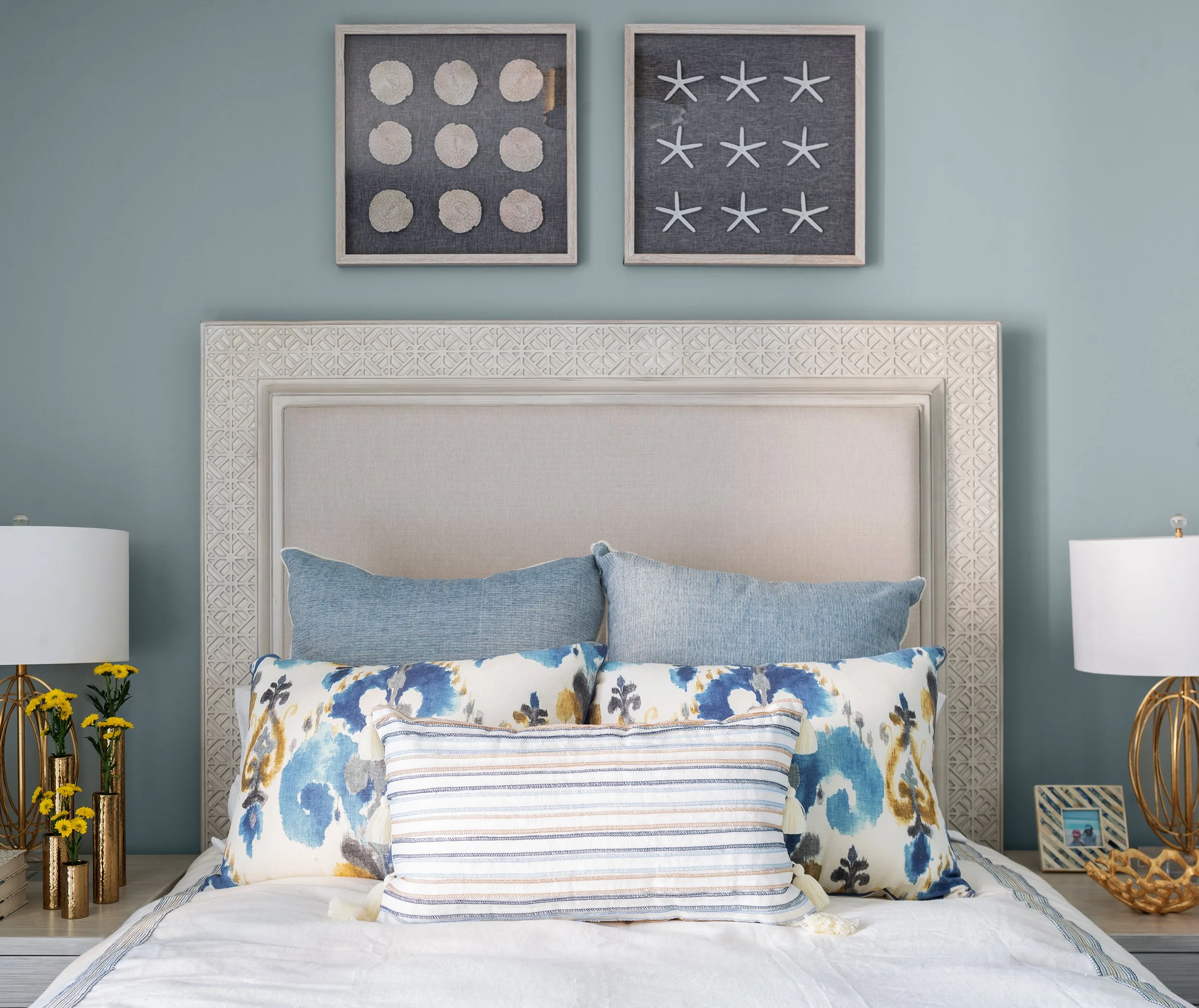 A bedroom with a beige upholstered headboard, blue pillows, and floral patterned accent pillows on a white bedspread; above the bed are two framed nautical themed artworks, one with white starfish and the other with white seashells; on each side of t