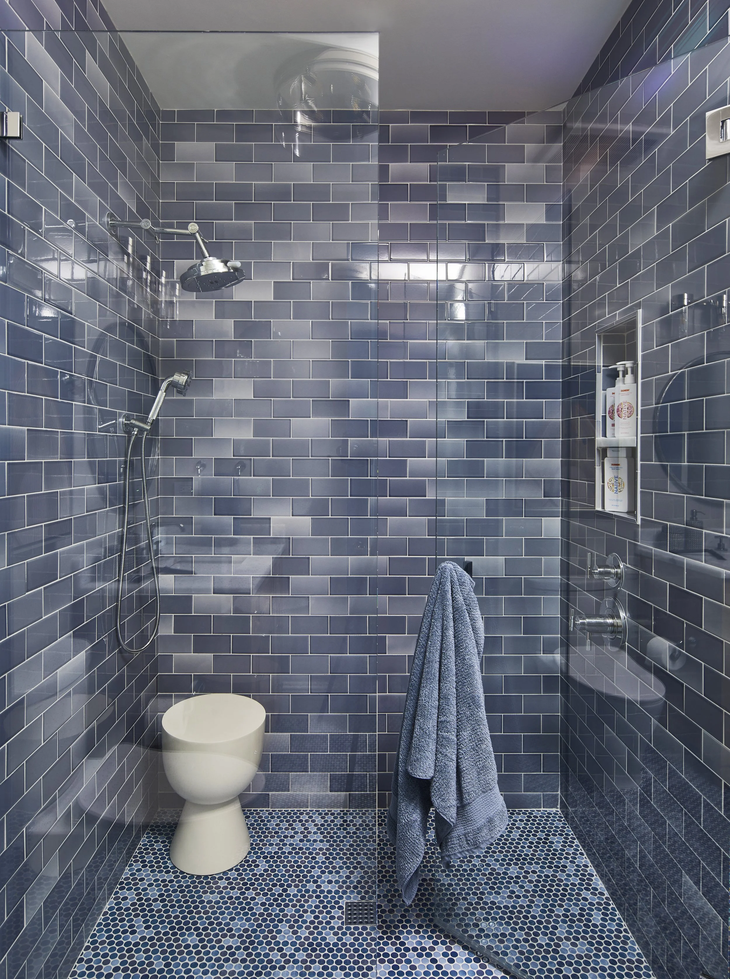 Bathroom with blue subway tile walls, a toilet, a showerhead, a handheld shower, a built-in shelf with toiletries, a towel hanging on a hook, and a glass shower door, with mosaic tile floor.