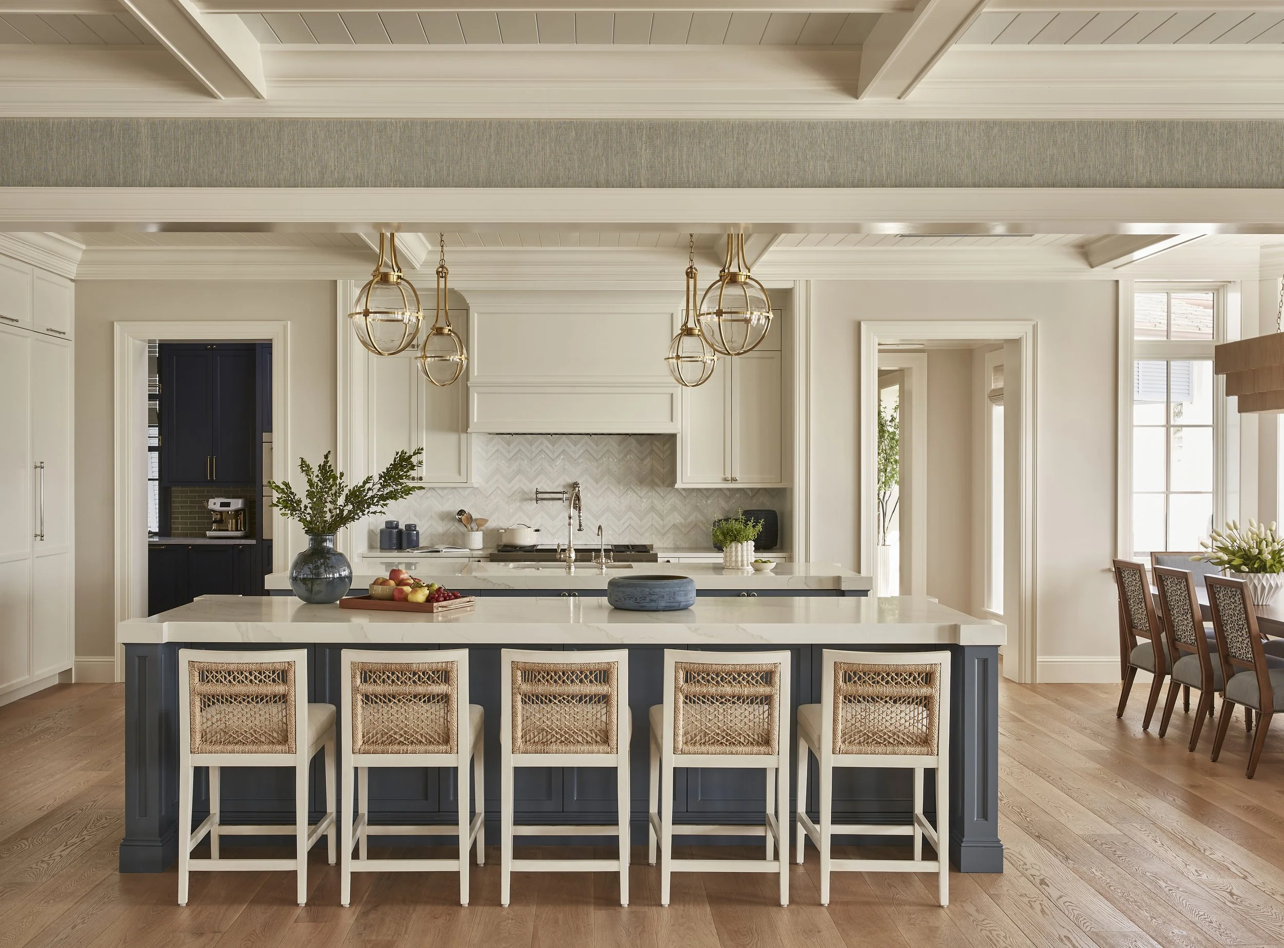 A spacious modern kitchen with a large island, navy blue base, and a marble countertop. Six woven rattan chairs with white frames are arranged around the island. White cabinetry, a herringbone backsplash, and gold pendant lights hang above the island