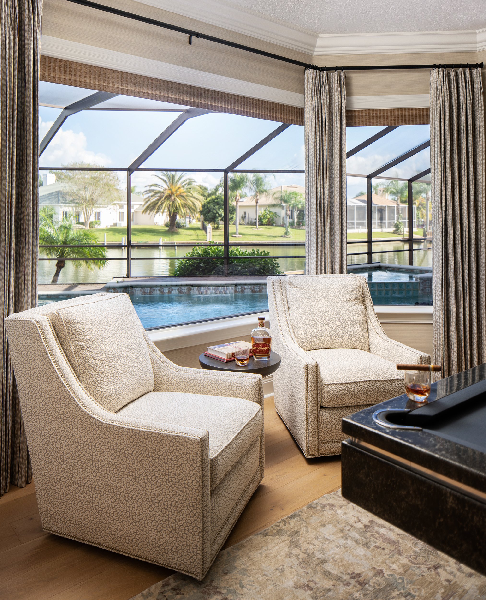 Living room with two armchairs, a small side table with whiskey bottles and glasses, large windows with a screened patio, water view, palm trees, and houses in the background.