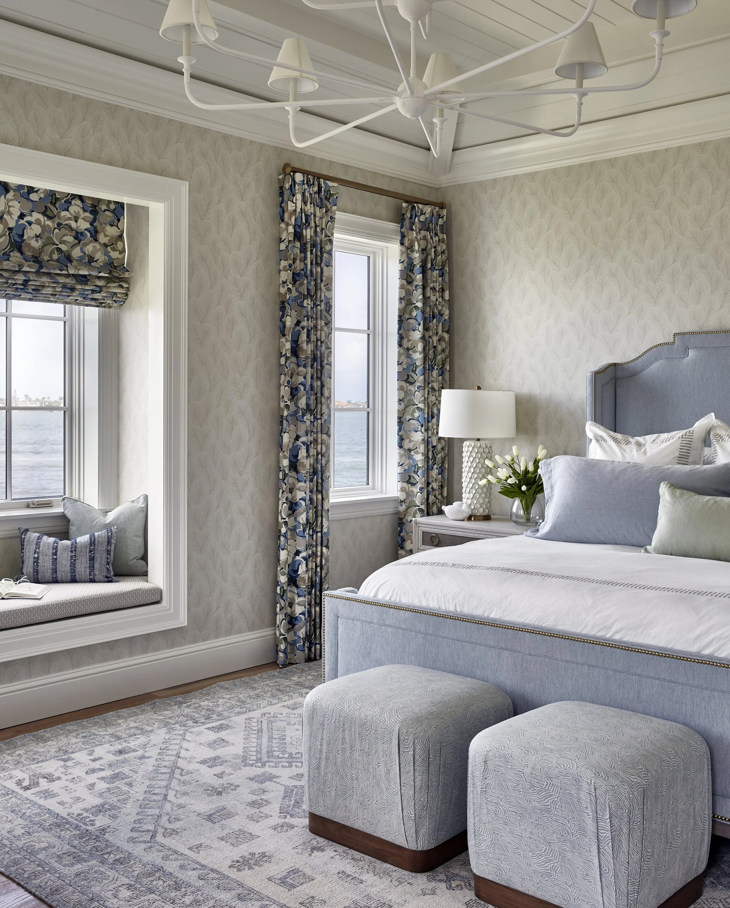 A bedroom with turquoise upholstered bed, multiple pillows, nightstand with white lamp, floral curtains, windows with view of water, and a window seat with pillows and a book.