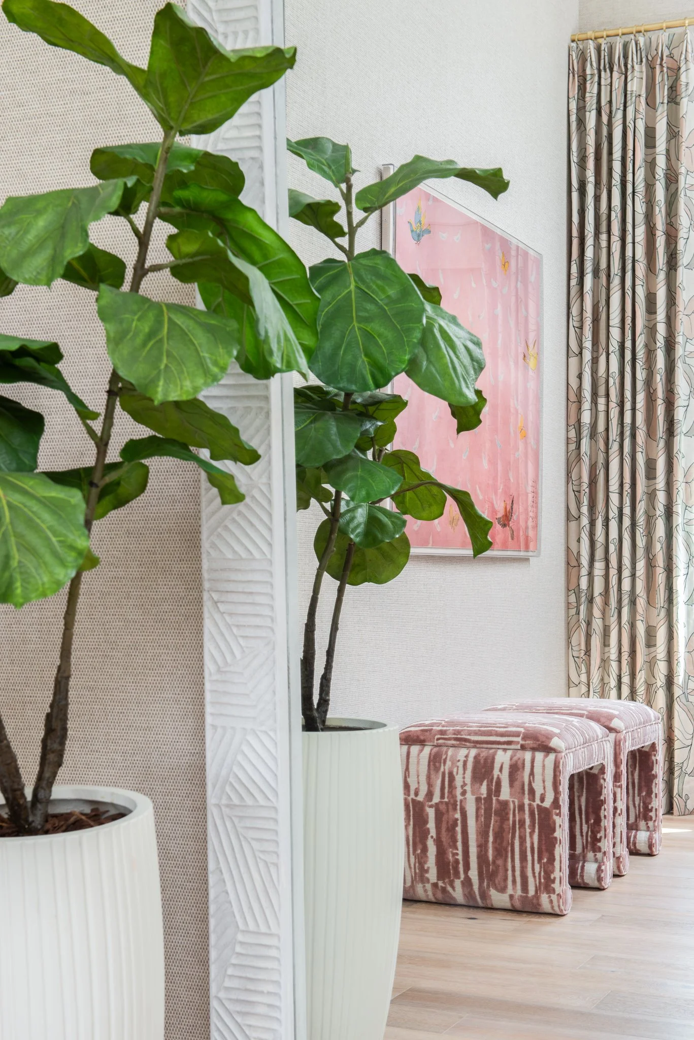 Interior space with large leafy potted plant, pink artwork with butterflies on wall, patterned drapes, and two pink striped upholstered stools.