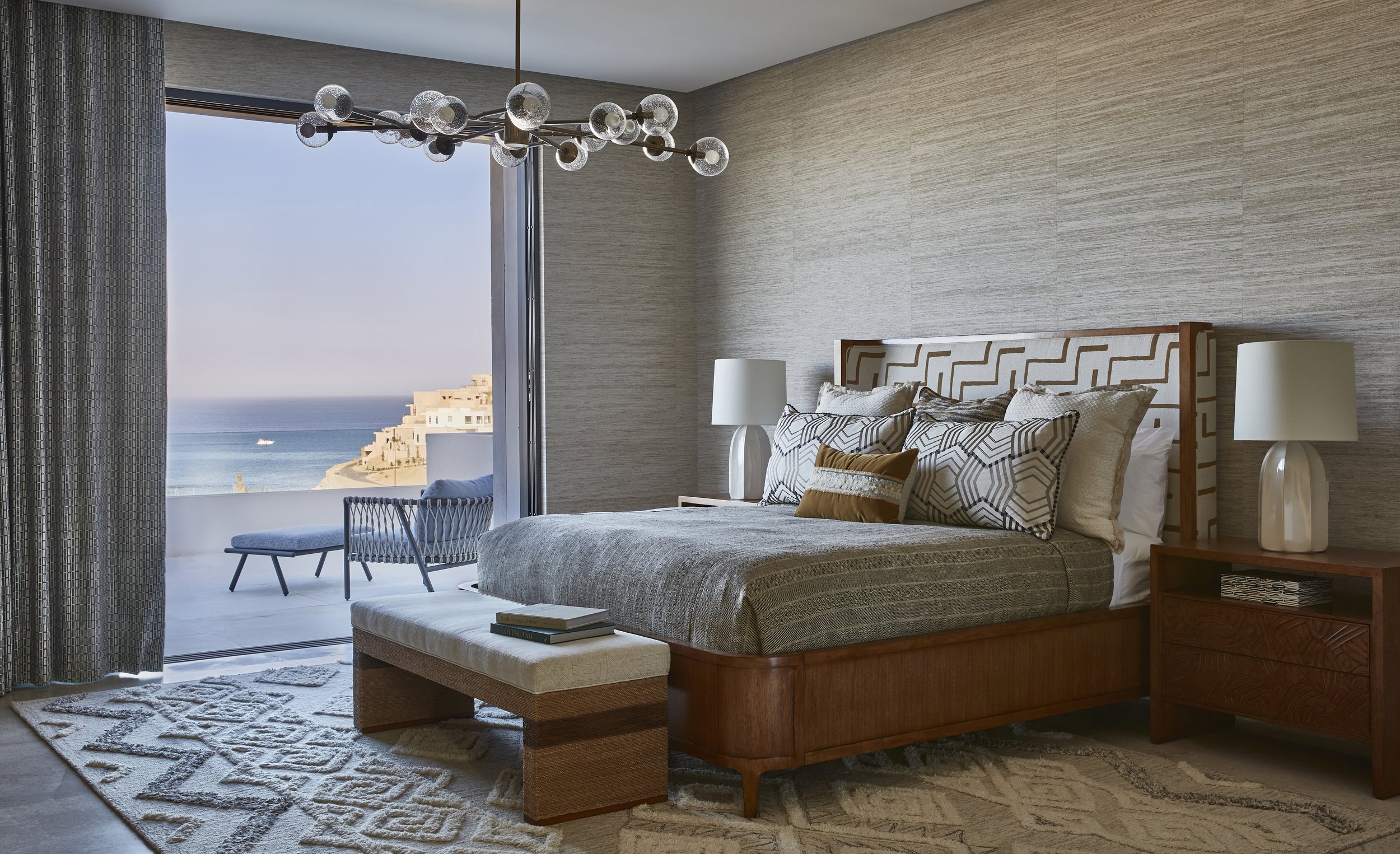 A bedroom with a view of the ocean through an open sliding glass door, showing outdoor chairs and a bench on a balcony. The room has a wooden bed frame, patterned pillows, bedside lamps, a rug, and a ceiling light fixture.