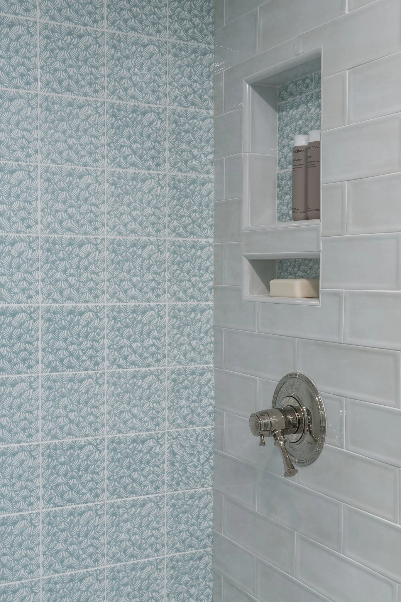 Close-up of a shower wall with white subway tiles and a built-in shelf, on which three bottles and a bar of soap are placed. The left side features decorative blue patterned tiles.