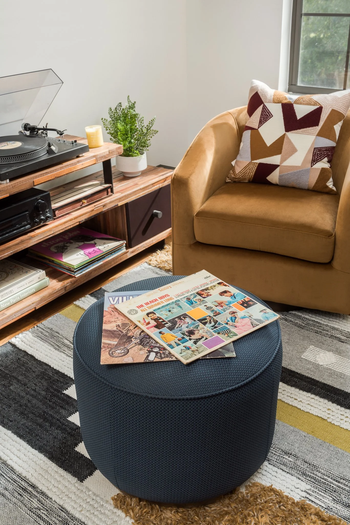 Living room with a record player on a wooden shelf, a yellow armchair with a colorful geometric pillow, a small round blue ottoman with vinyl records on top, a potted plant, and a window with greenery outside.