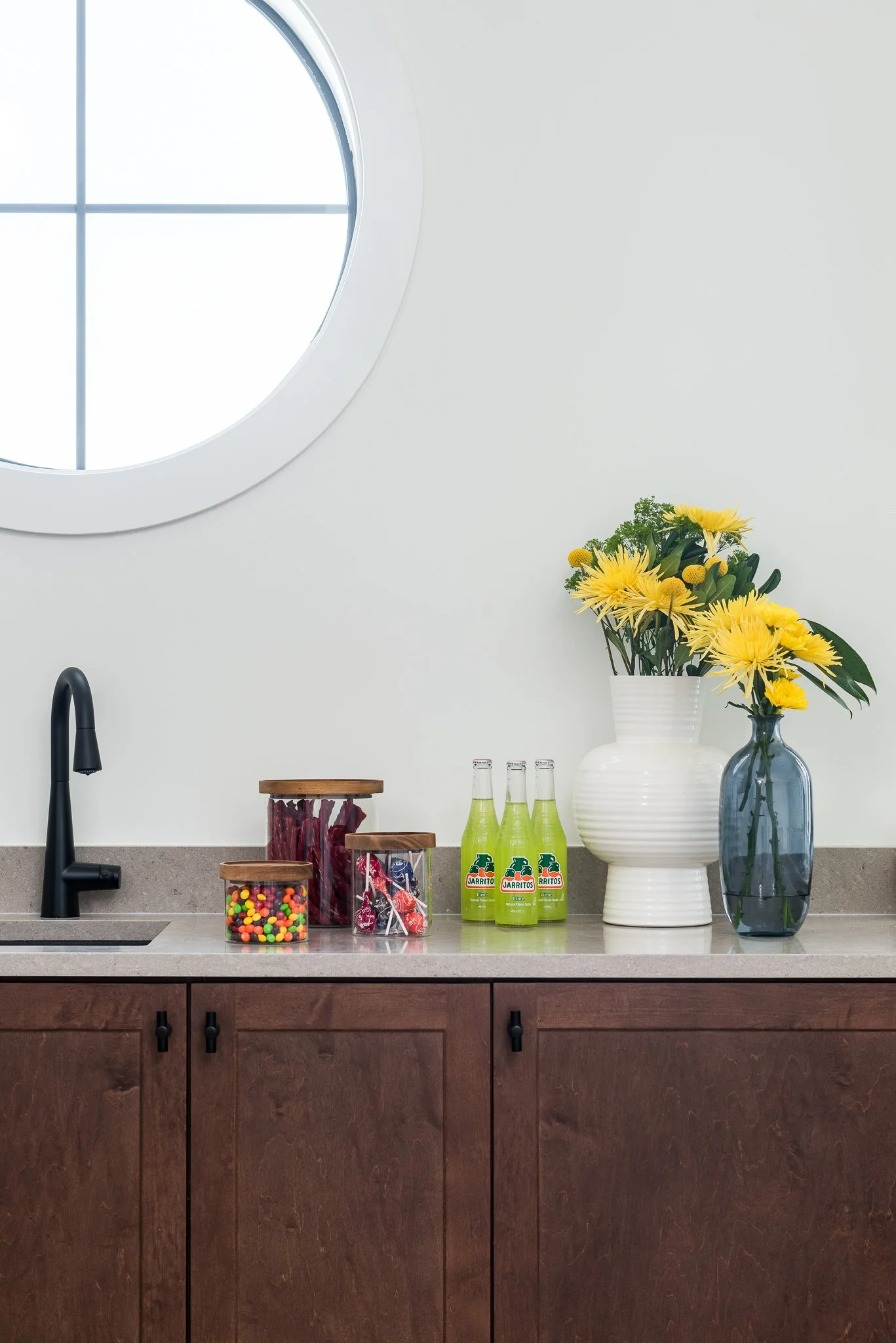 Kitchen countertop with black faucet, three jars with candies, three bottles of green jarritos soda, a large white vase with yellow flowers, a smaller dark blue vase with yellow flowers, and a round window in the white wall above the counter.