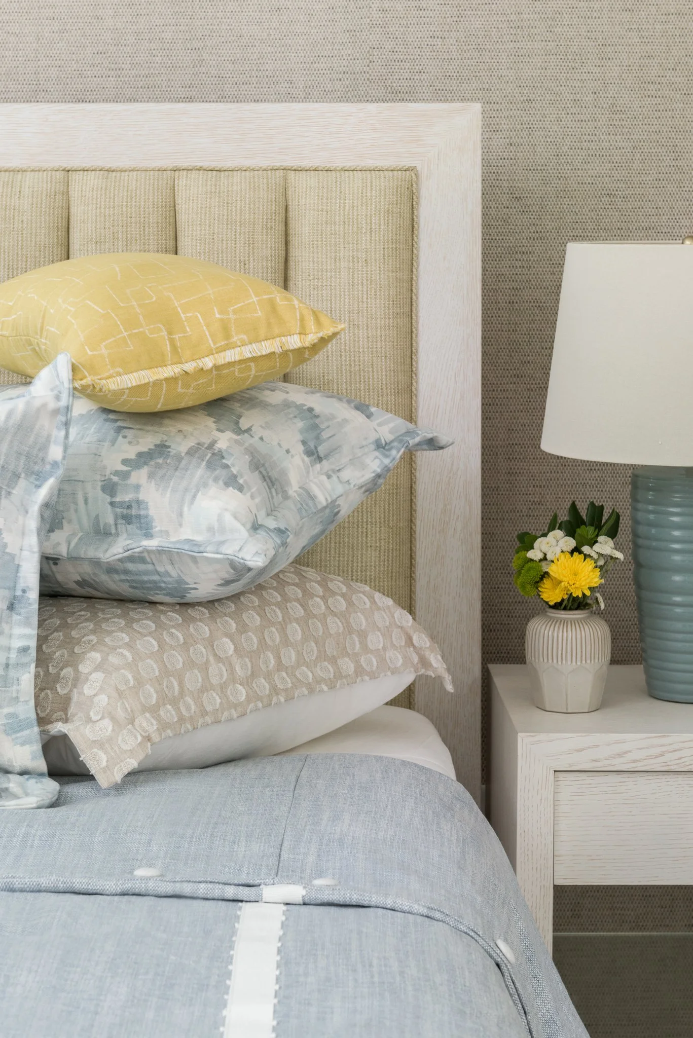 Close-up of a bed with stacked pillows and a nightstand with a vase of flowers and a lamp.