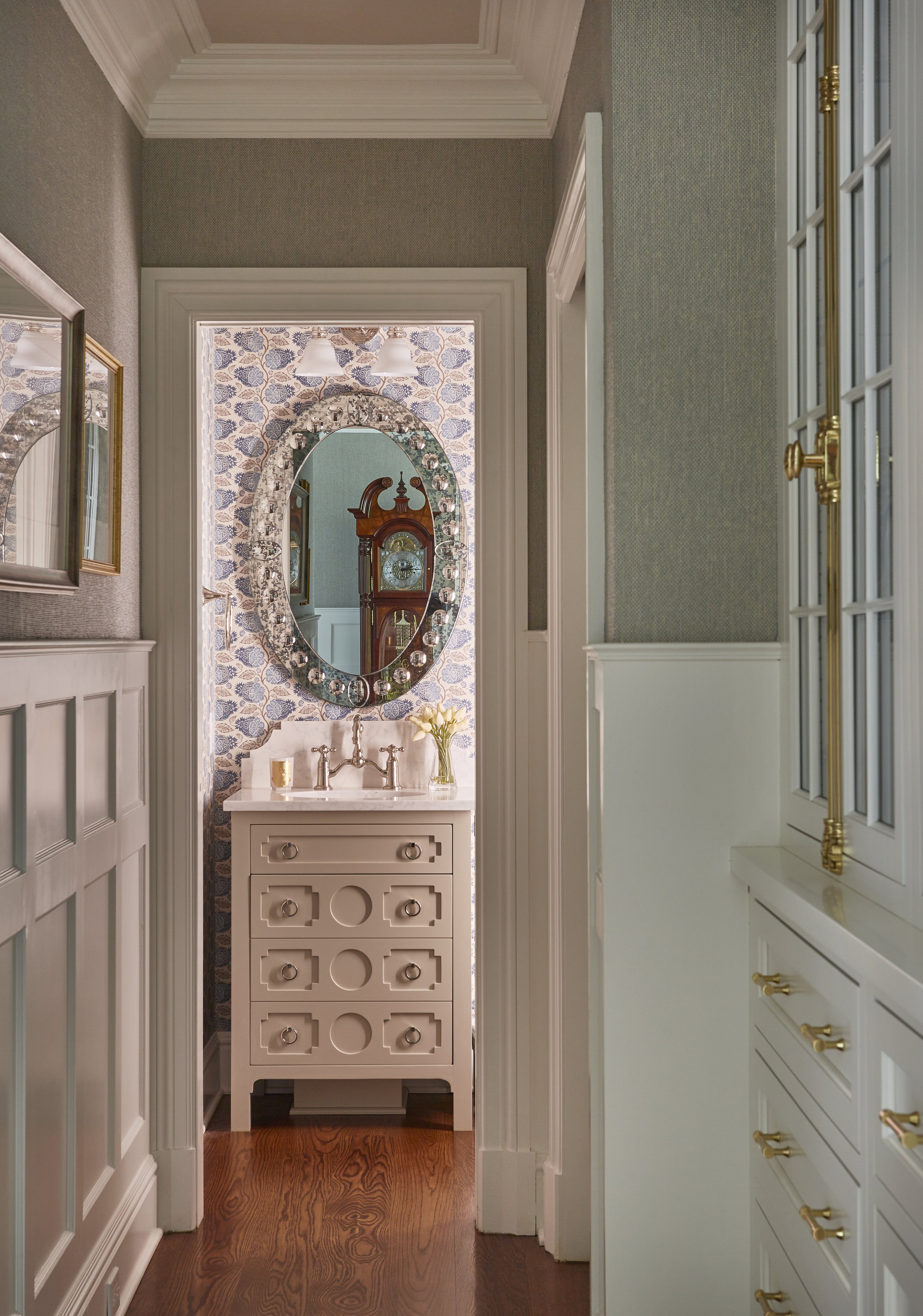 View of a small bathroom with an oval mirror, floral wallpaper, a white vanity, and tall cabinets with gold handles, hardwood flooring.