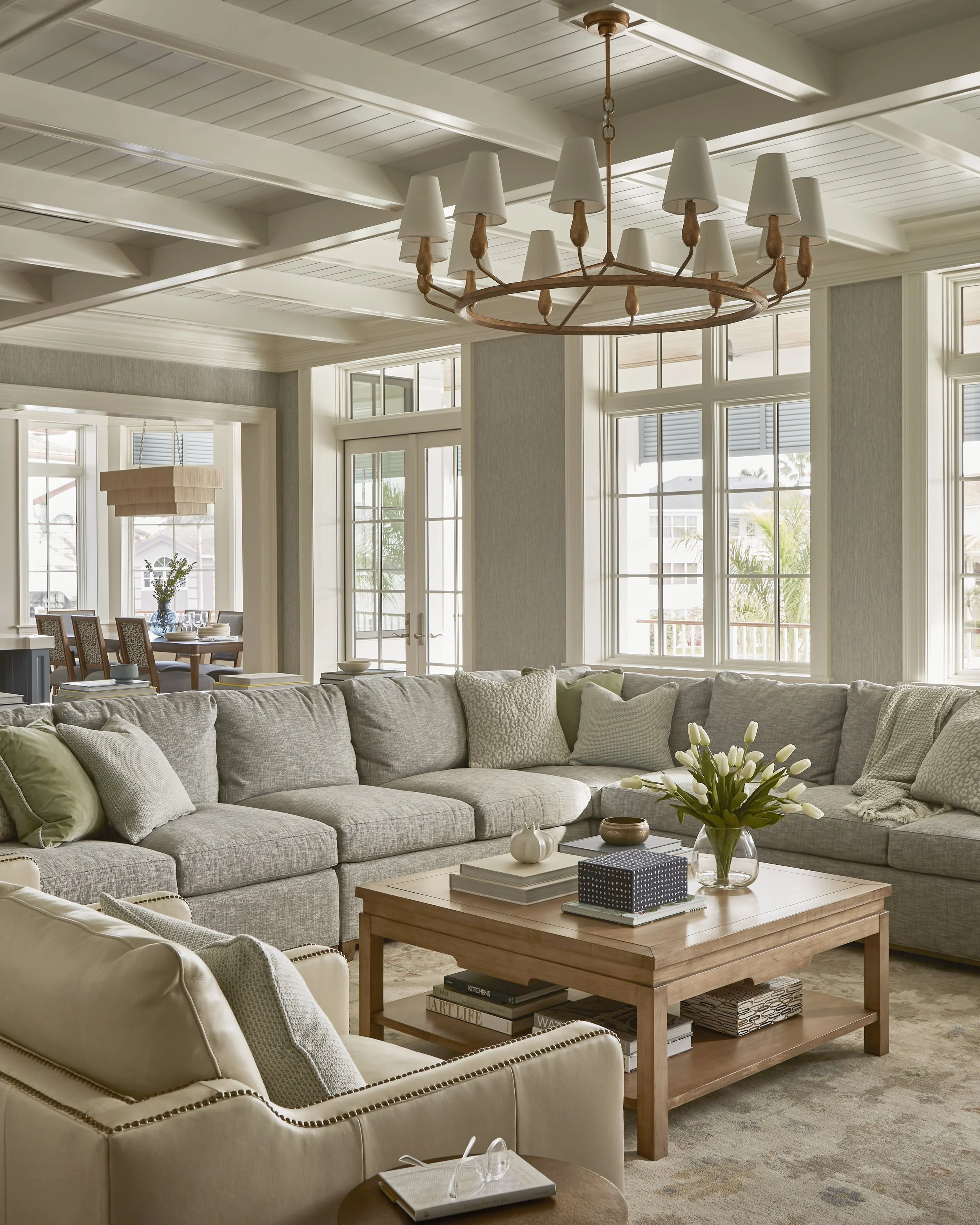 Sunlit living room with a large sectional sofa, a wooden coffee table with decorative items and books, and multiple large windows with outside views.