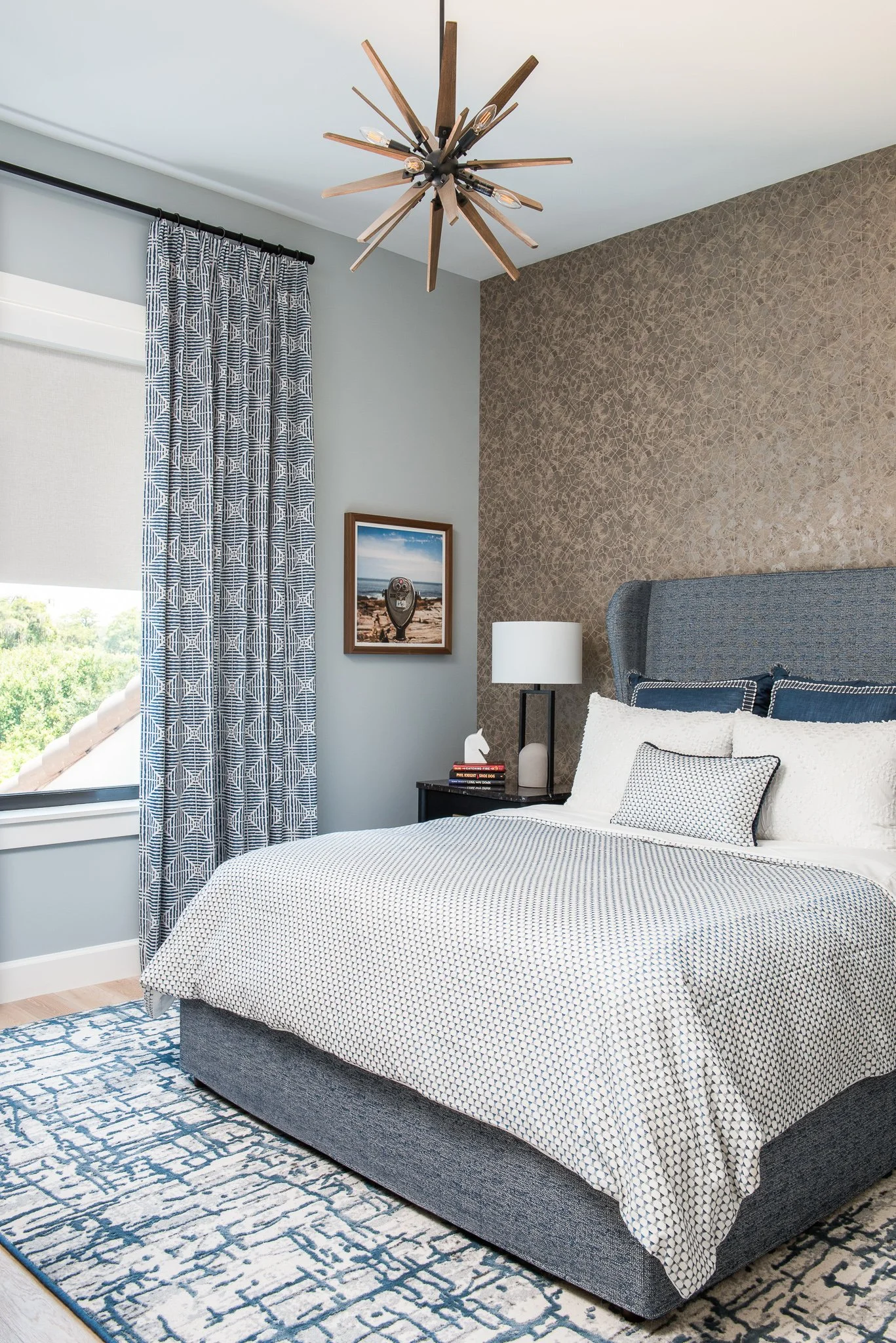 A bedroom with a white and black patterned bed, blue and white pillows, a textured wall behind the bed, a nightstand with a lamp, books, and decorative items, a window with patterned blue curtains, and a unique wooden light fixture on the ceiling.