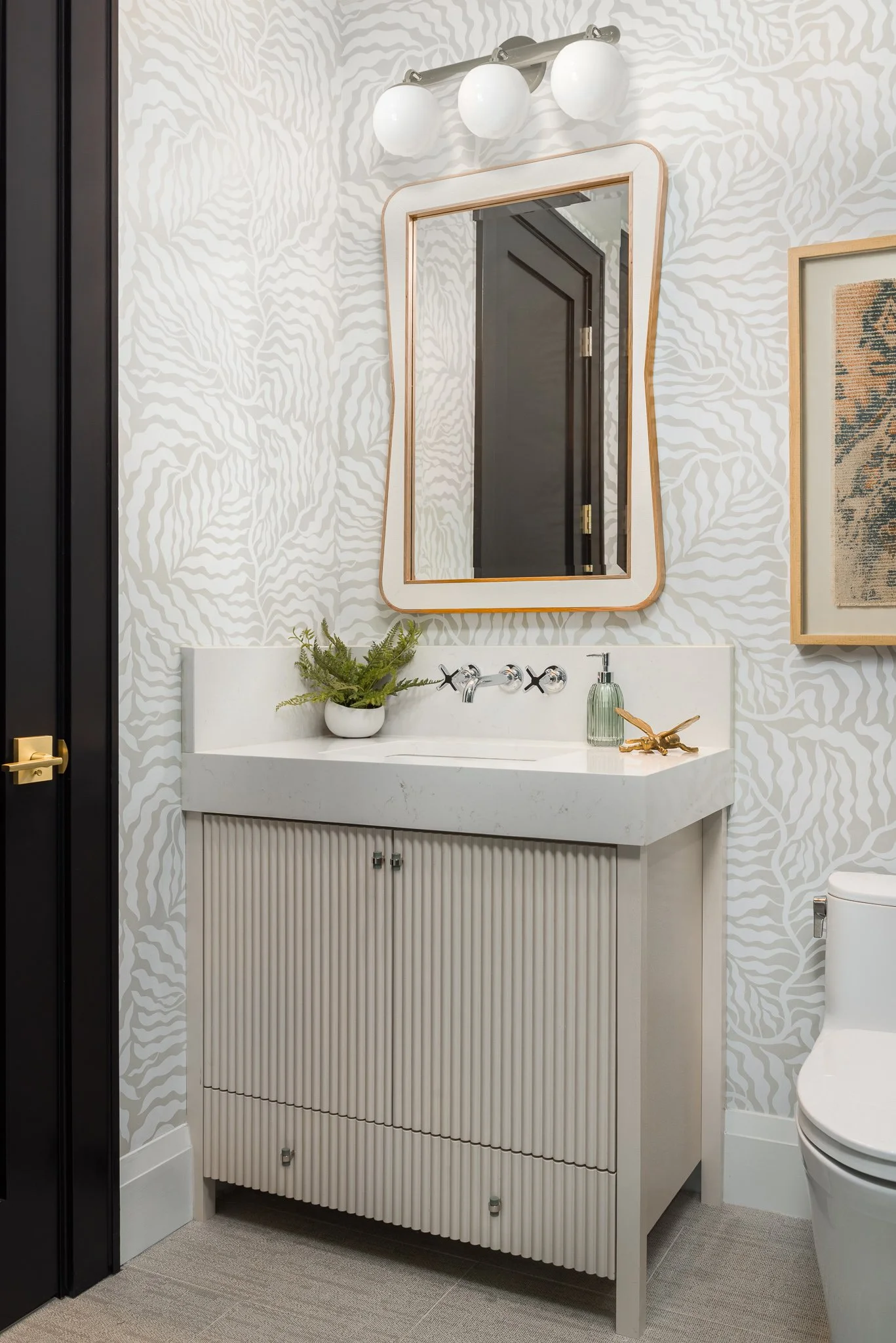 Bathroom with patterned wallpaper, white vanity with ribbed cabinet doors, a large mirror, a plant, soap dispenser, and chrome fixtures.