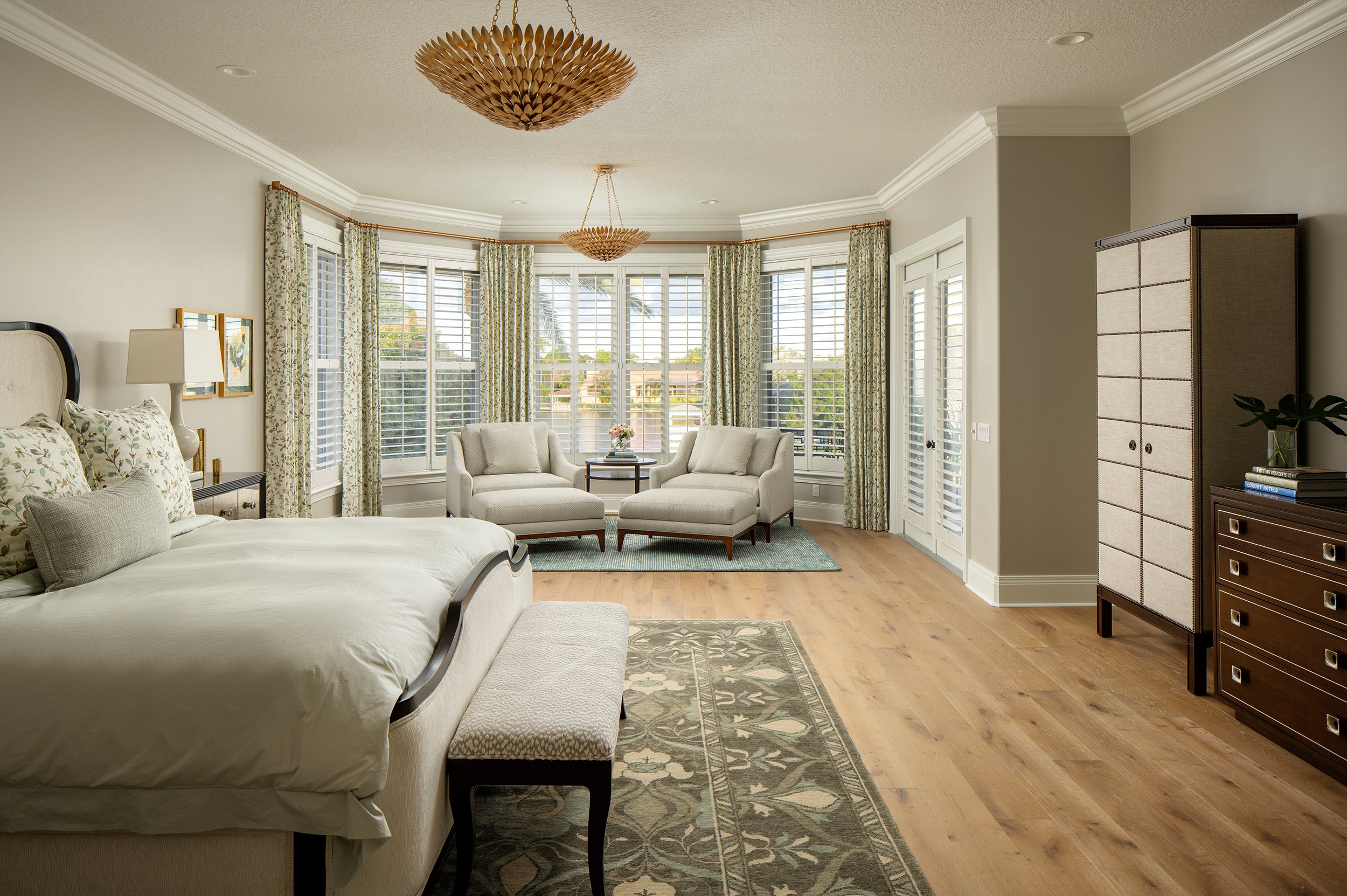 Bright bedroom with a large bay window, seating area with two armchairs, a small table with flowers, a bed with pillows, patterned curtains, hardwood floor, and a wardrobe.