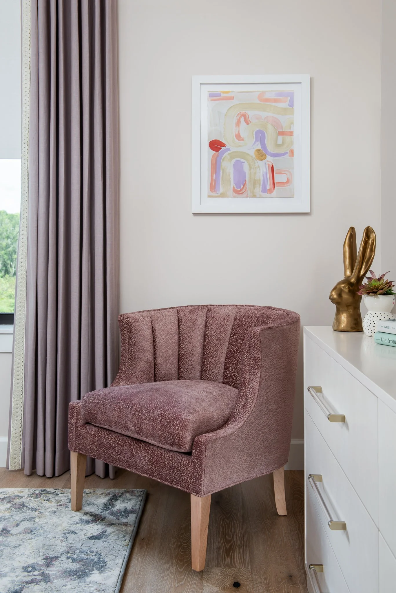 A pink textured armchair next to a white dresser with gold handles, a gold bunny sculpture, potted plant, and books, with a window and light-colored curtain in the background.