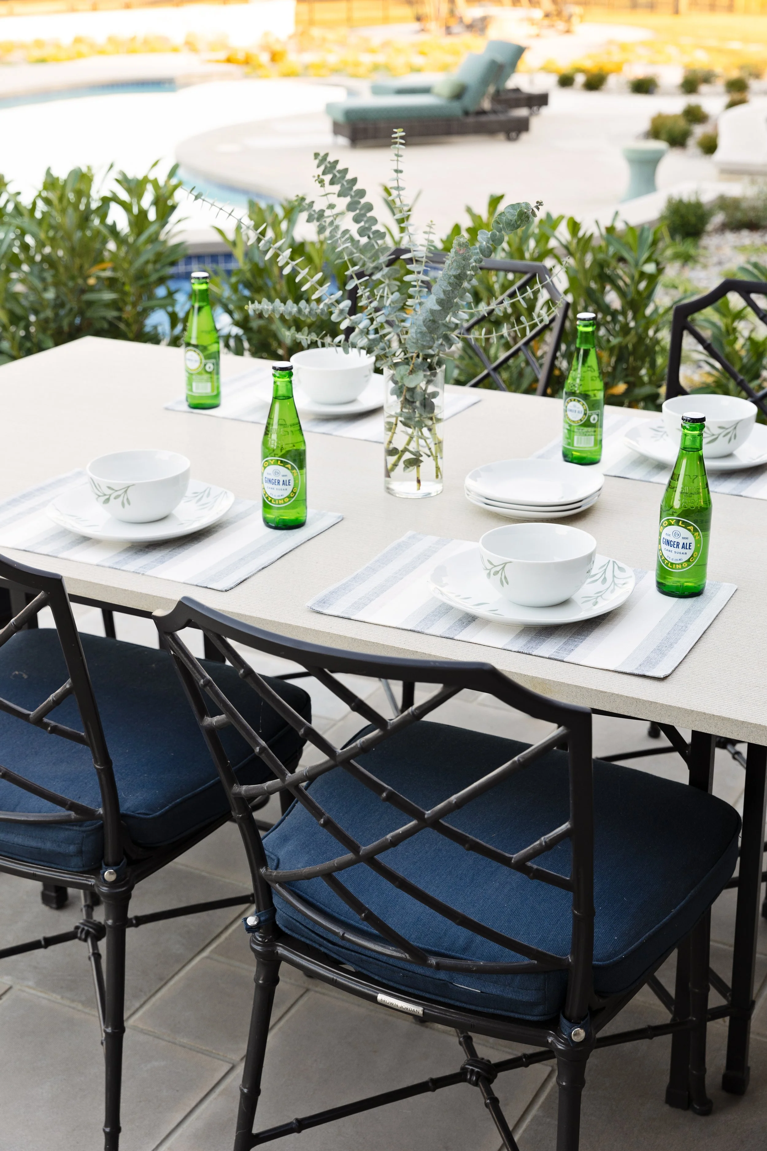 Outdoor dining table set with white plates, bowls, green glass bottles of ginger ale, and a centerpiece of eucalyptus in a glass vase, with a swimming pool and lounge chairs in the background.