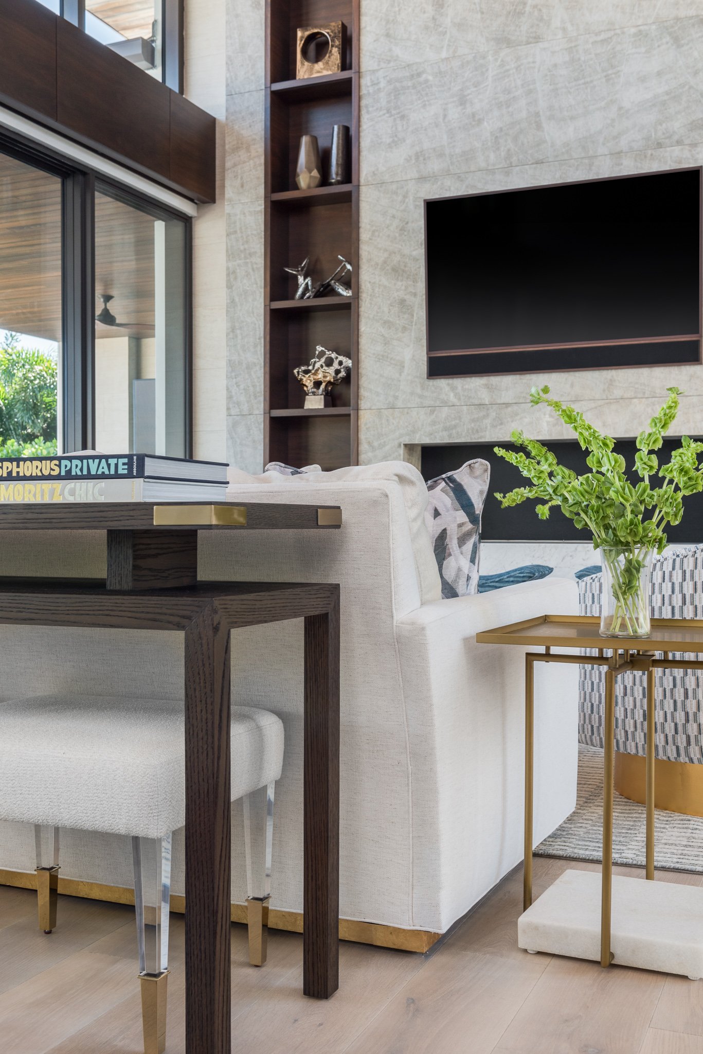 Modern living room with a white sofa, a gold side table with a vase of green plants, a flat-screen TV on the wall, and a wooden bookshelf with decorative objects.