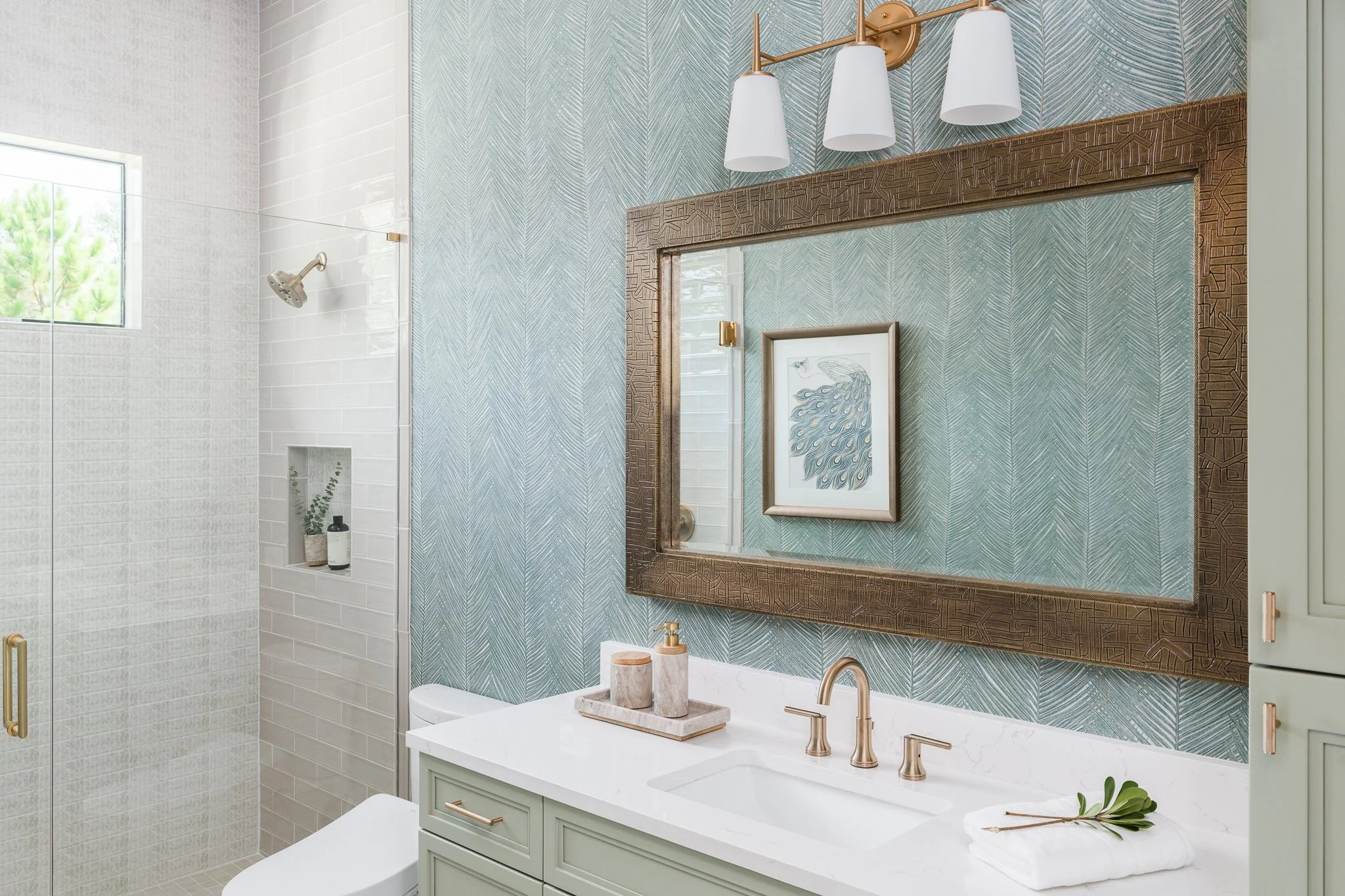 Modern bathroom with a white vanity, gold fixtures, and a large decorative frame mirror. The shower area has white subway tiles and a small window, with a niche holding toiletries. The wall behind the mirror has textured blue wallpaper with a framed 