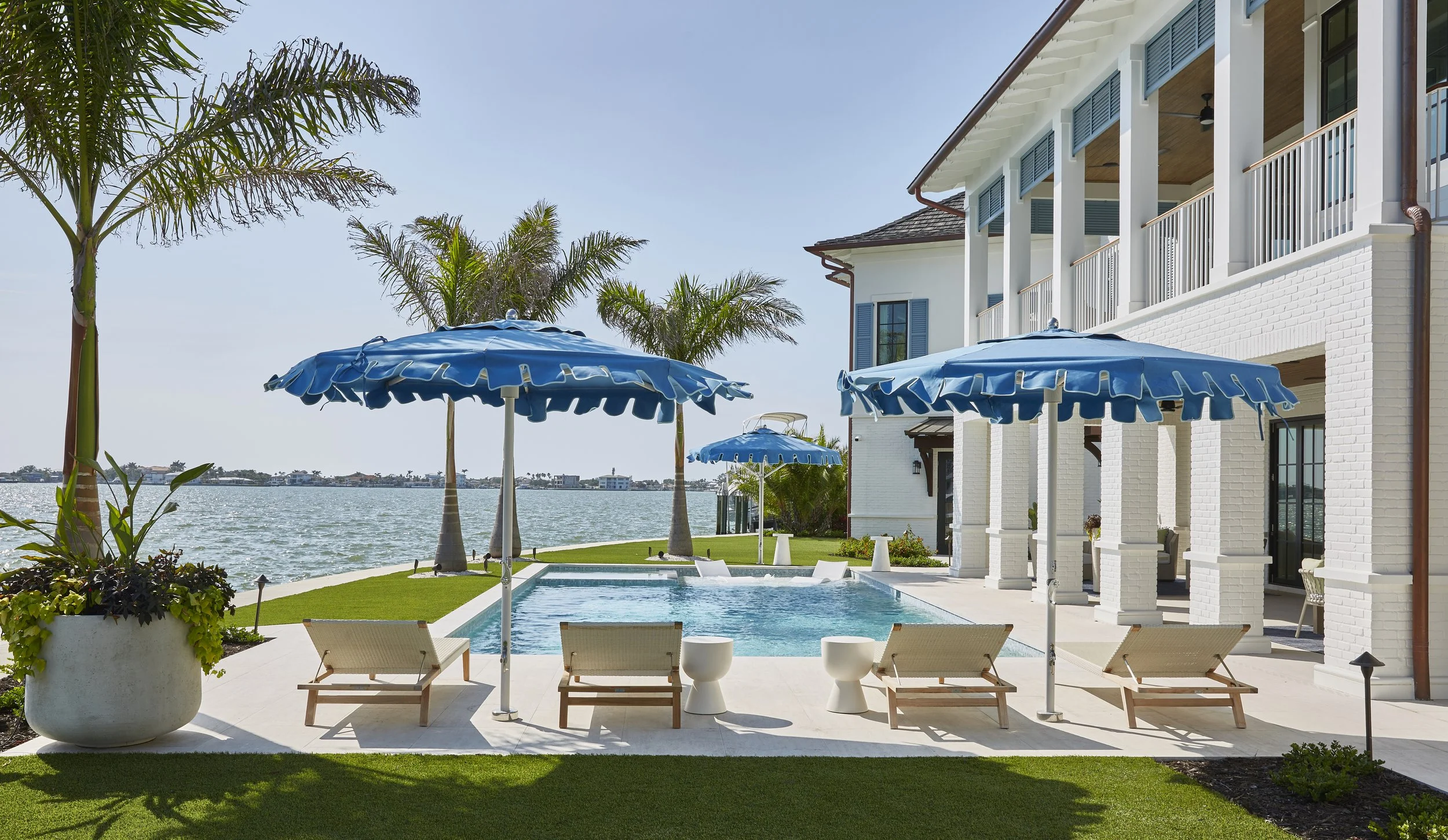 Backyard view of a lake with a pool, four lounge chairs, three blue umbrellas, palm trees, and a white house with a balcony.