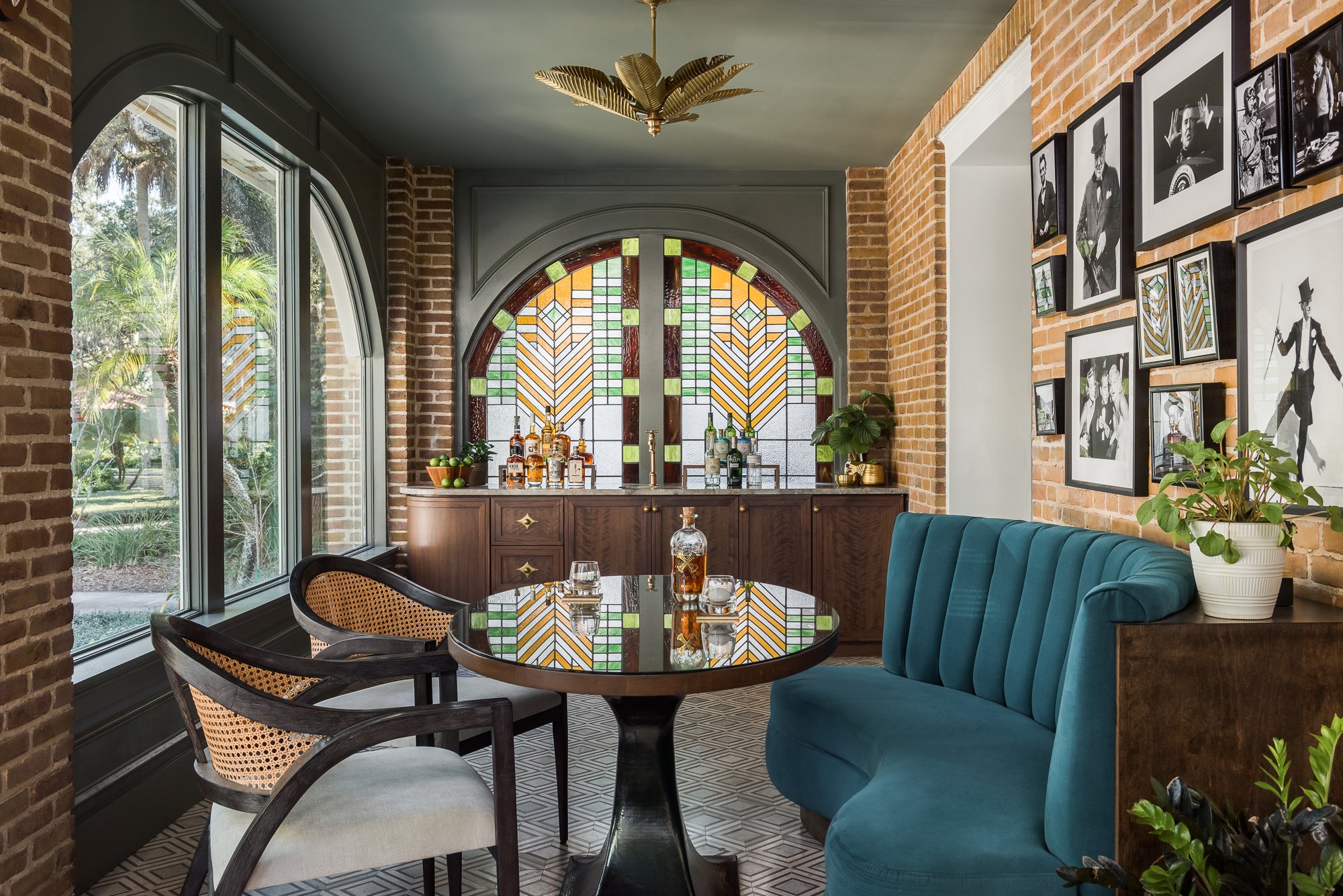 Interior of a stylish living or dining area with exposed brick walls, stained glass windows, a teal tufted bench, a wooden dining table with glassware, bar setup with liquor bottles, framed black-and-white photos on the wall, and decorative plants.