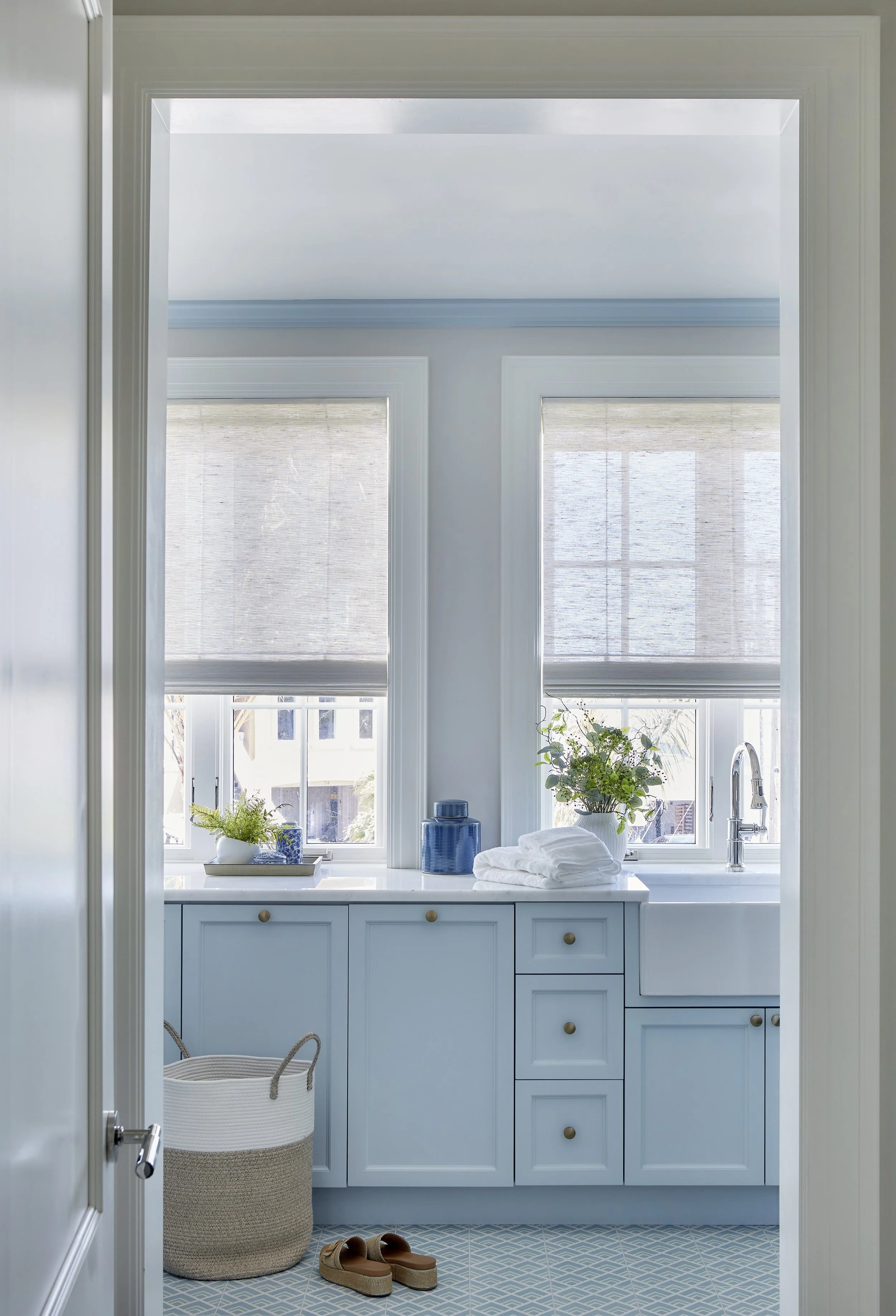 Bright bathroom with light blue cabinets, two windows with shades, plants, and folded towels on the sink countertop.