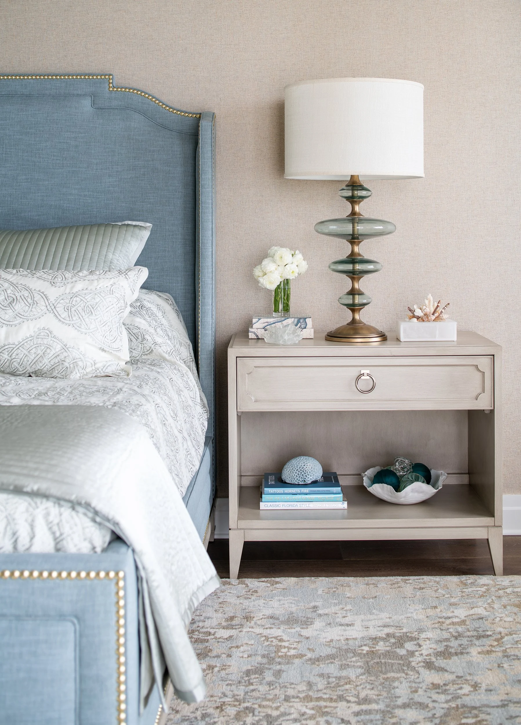A bedroom with a large blue upholstered bed, white patterned bedding, a beige bedside table with a seashell decor, a lamp, a vase of white flowers, and books, set against a beige wall with a patterned rug on the floor.