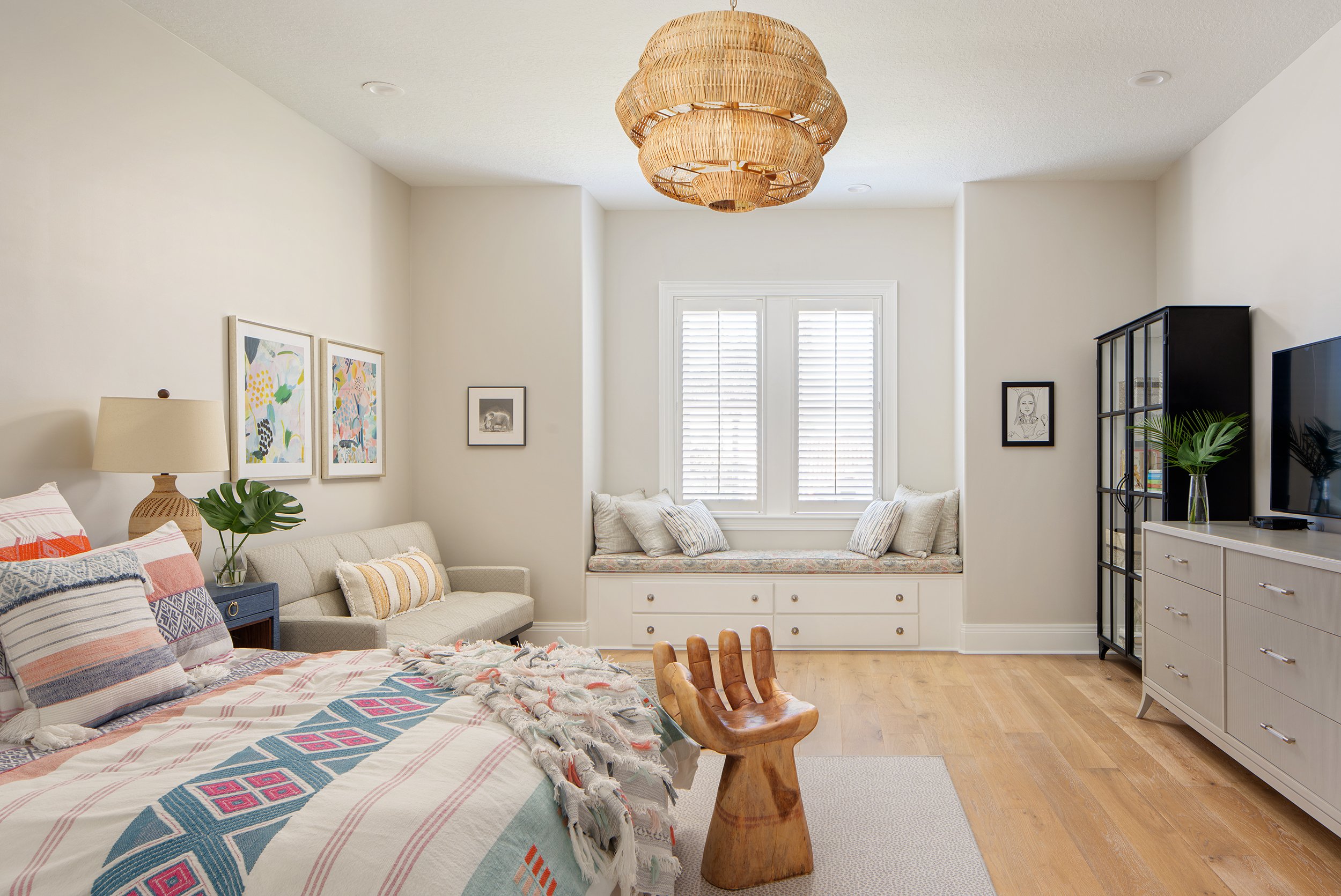 A bright, modern bedroom with a window seat, artwork, and various furniture including a bed, a chair, a dresser, and a black shelving unit, all decorated with plants and colorful accents.