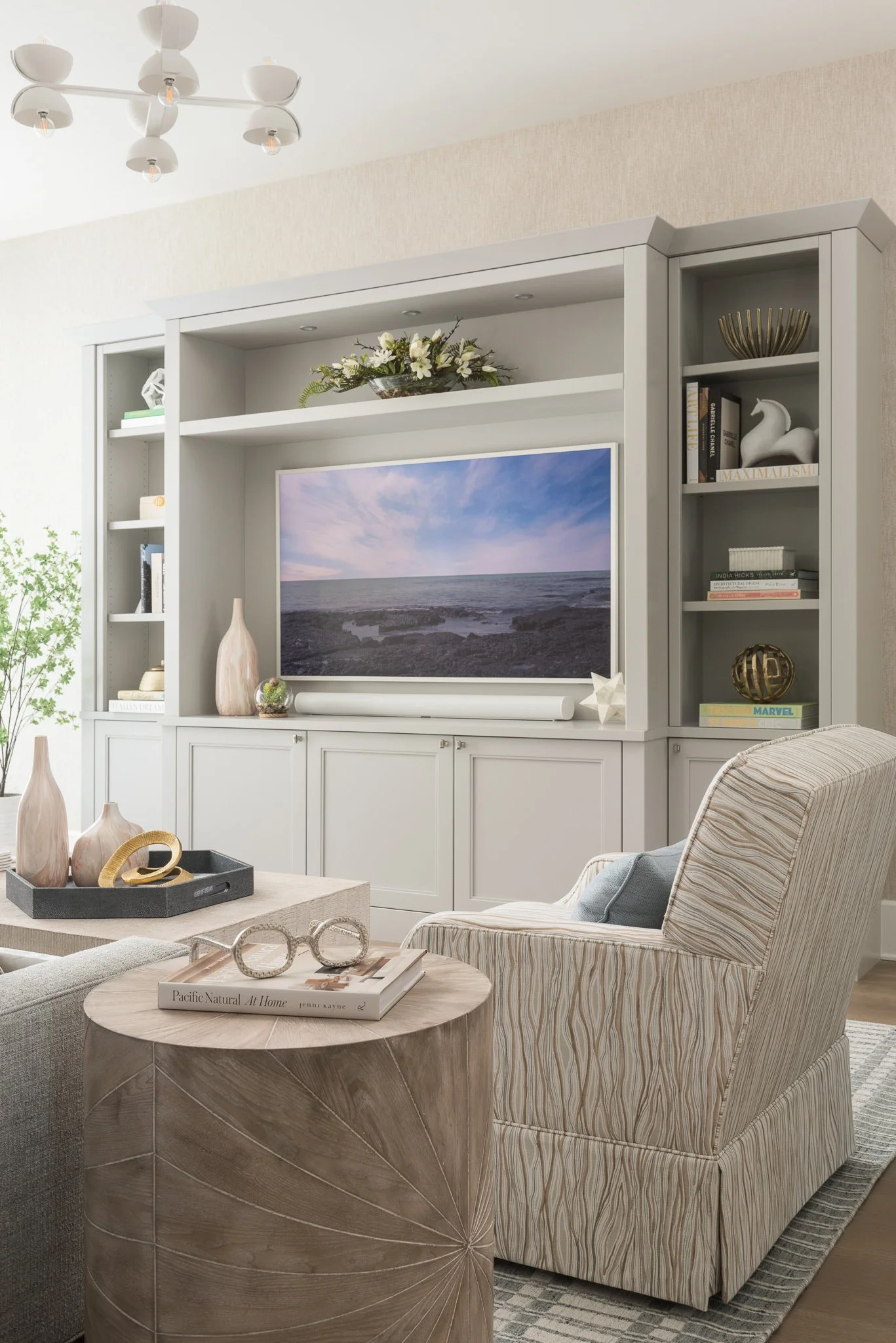 Living room with a large entertainment center containing a flat screen TV, decorative vases, books, and ornaments. A striped upholstered armchair is beside a round wooden side table holding glasses and a book. A modern ceiling light fixture is overhe