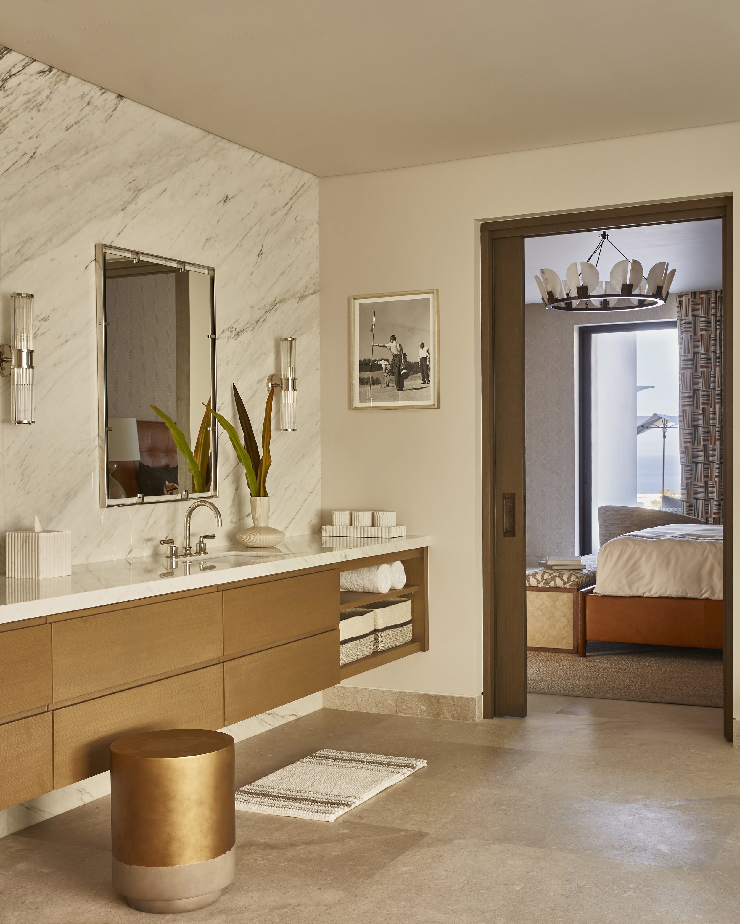 Luxurious hotel bathroom with a marble wall, wooden vanity, mirror, plants, and towels, leading to a bedroom with a bed, chairs, and ocean view.