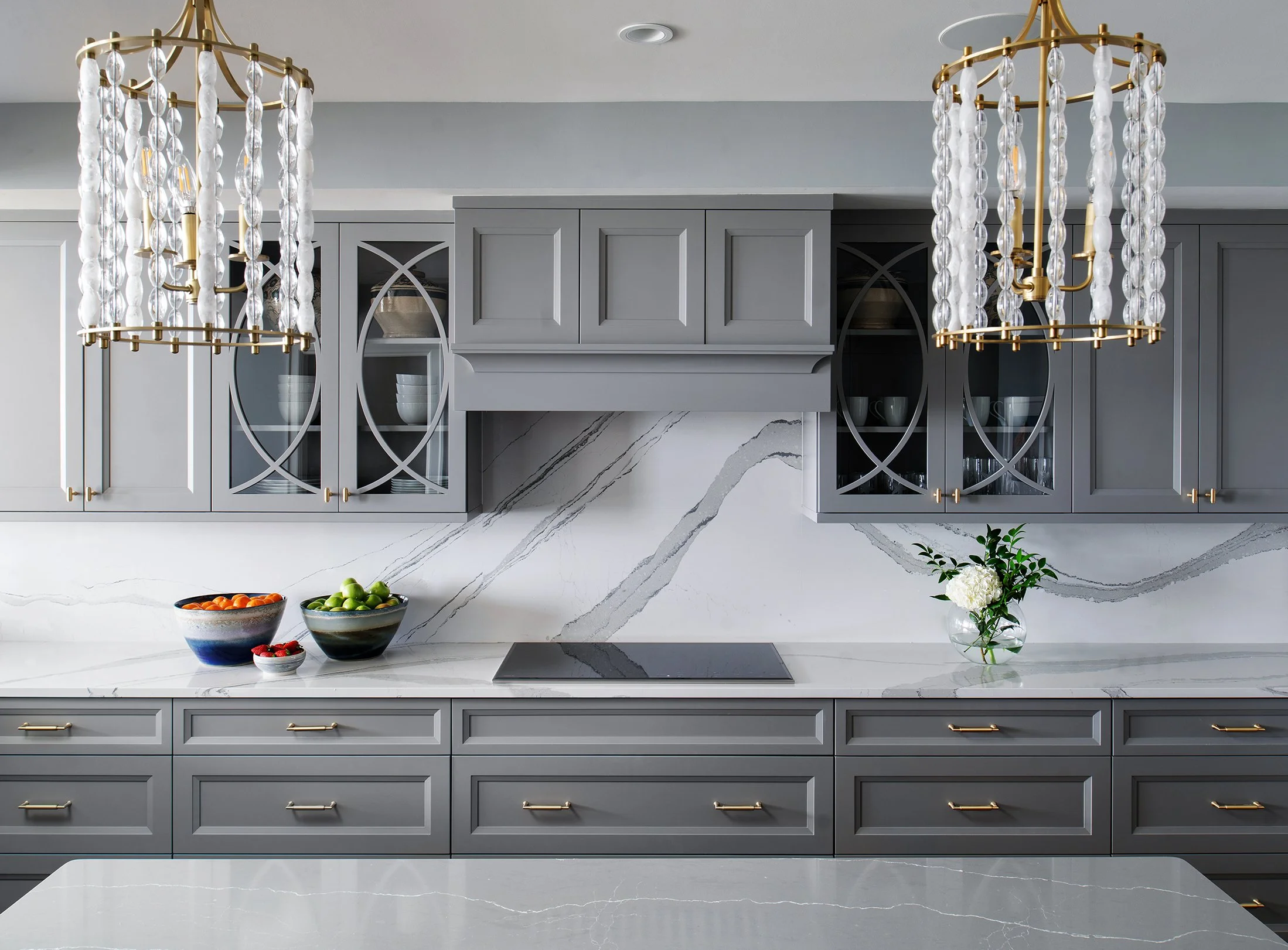 Modern kitchen with gray cabinetry, white marble countertops and backsplash, two gold chandeliers with hanging crystals, and a glass vase with white flowers.