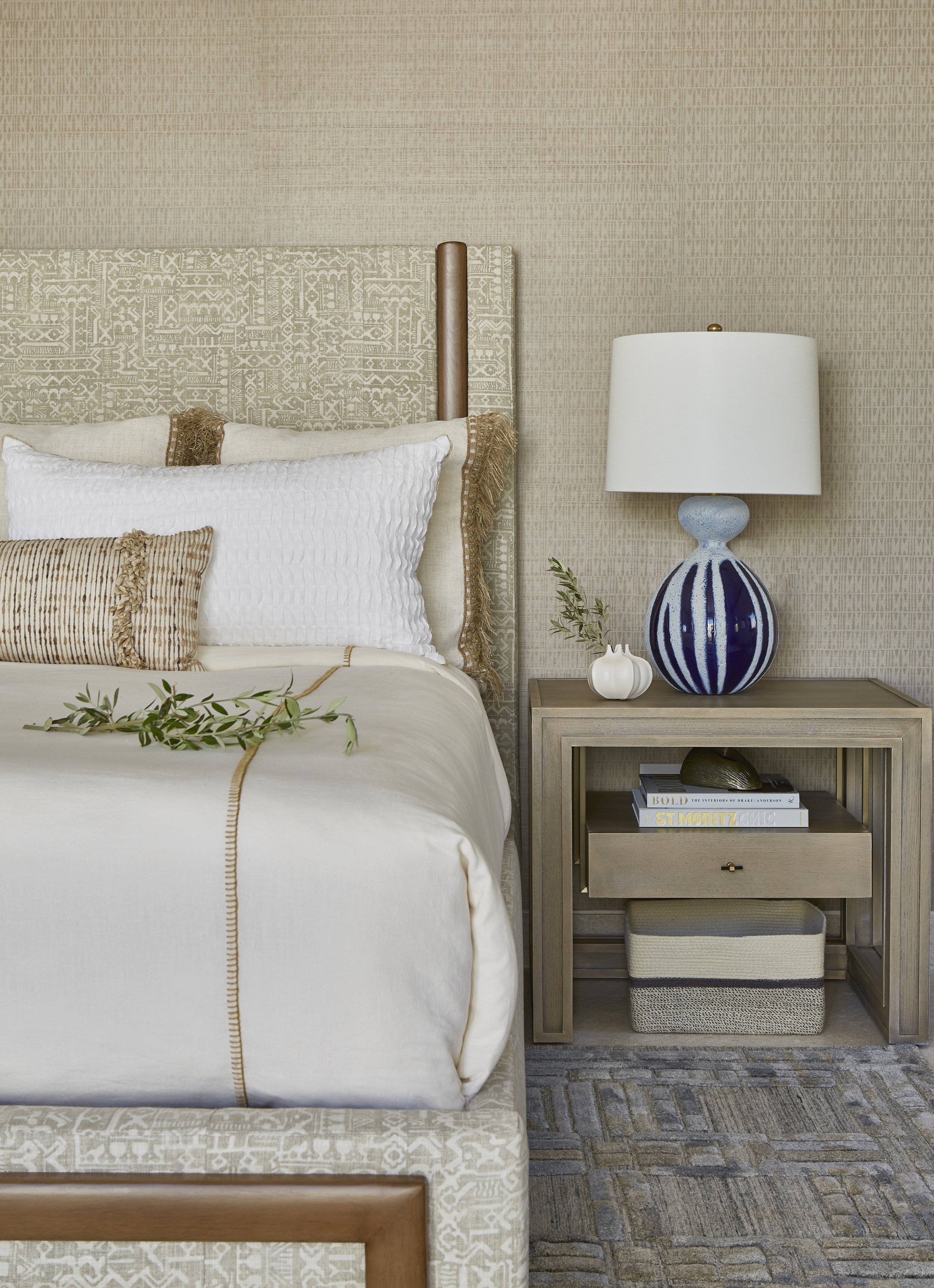 A bedroom with a bed, pillows, and blankets, a nightstand with a blue and white lamp, vases, books, and a woven basket, and a textured beige wall and carpet.