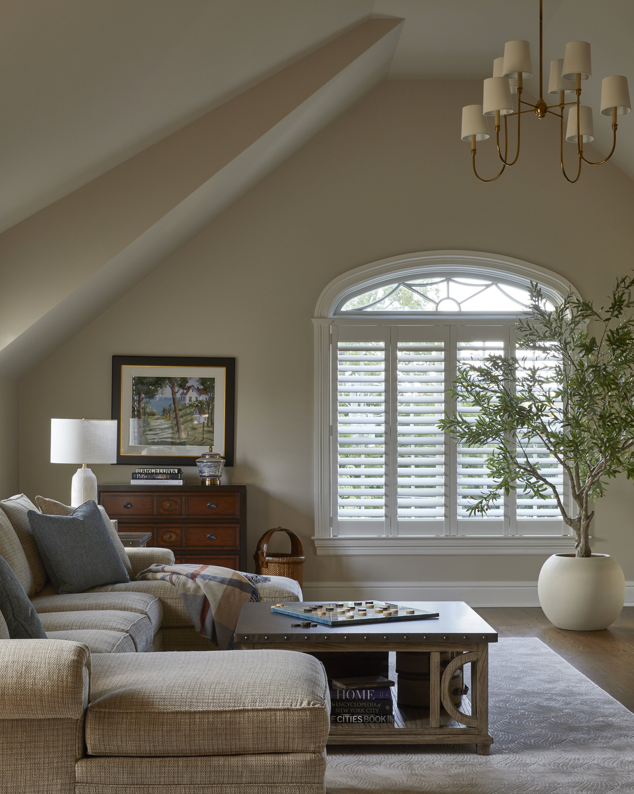 Living room with beige sofa, wooden coffee table, large window with white shutters, potted plant, and chandelier.