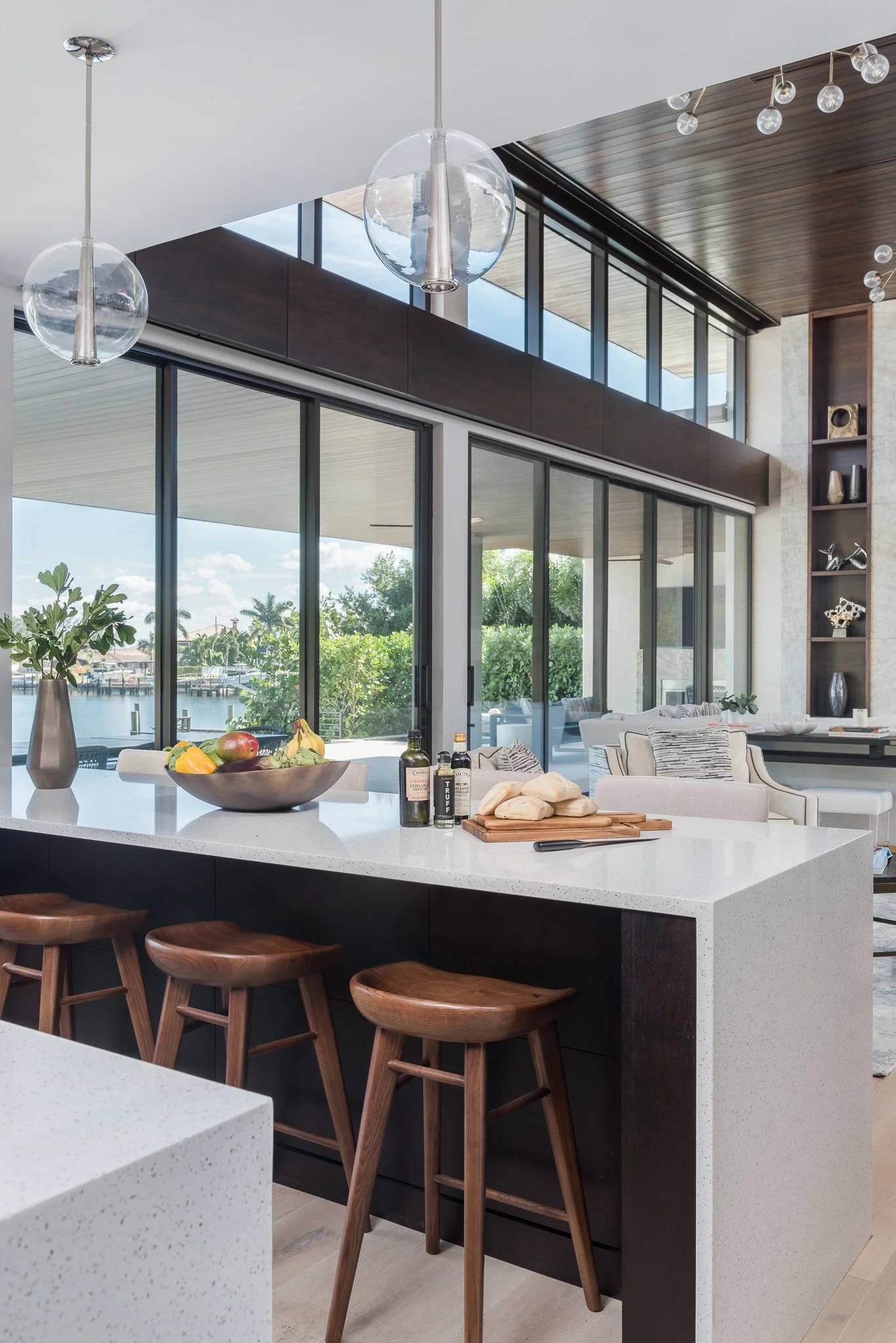 Modern open-concept kitchen and living room with large windows overlooking a waterfront view, featuring a white island with a bowl of fruit, wooden bar stools, and contemporary decor.