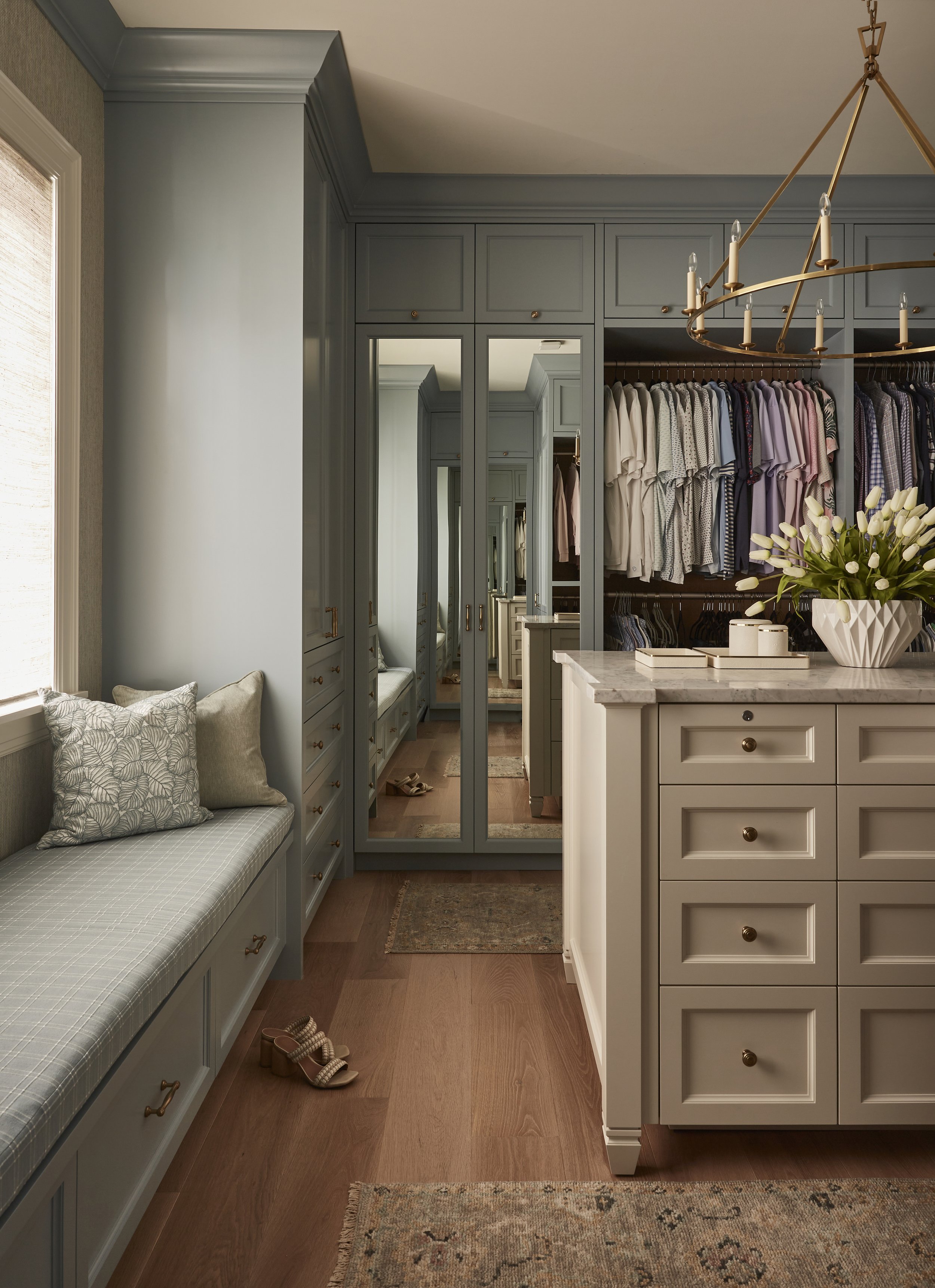 A walk-in closet with built-in wardrobes and drawers, a marble-topped island, a window seat with cushions, and a pair of heels on the floor.