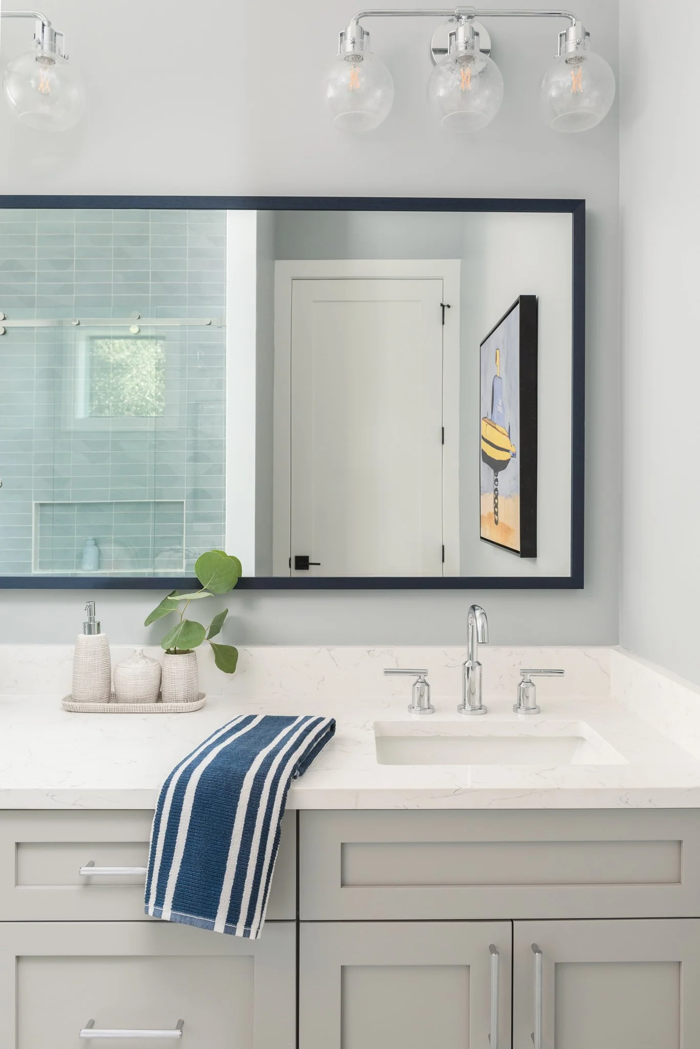 Modern bathroom vanity with a large mirror, white marble countertop, and light gray cabinets. There are two beige soap dispensers and a small plant on a tray, a blue and white striped towel draped over the counter, and a view of a shower area with li