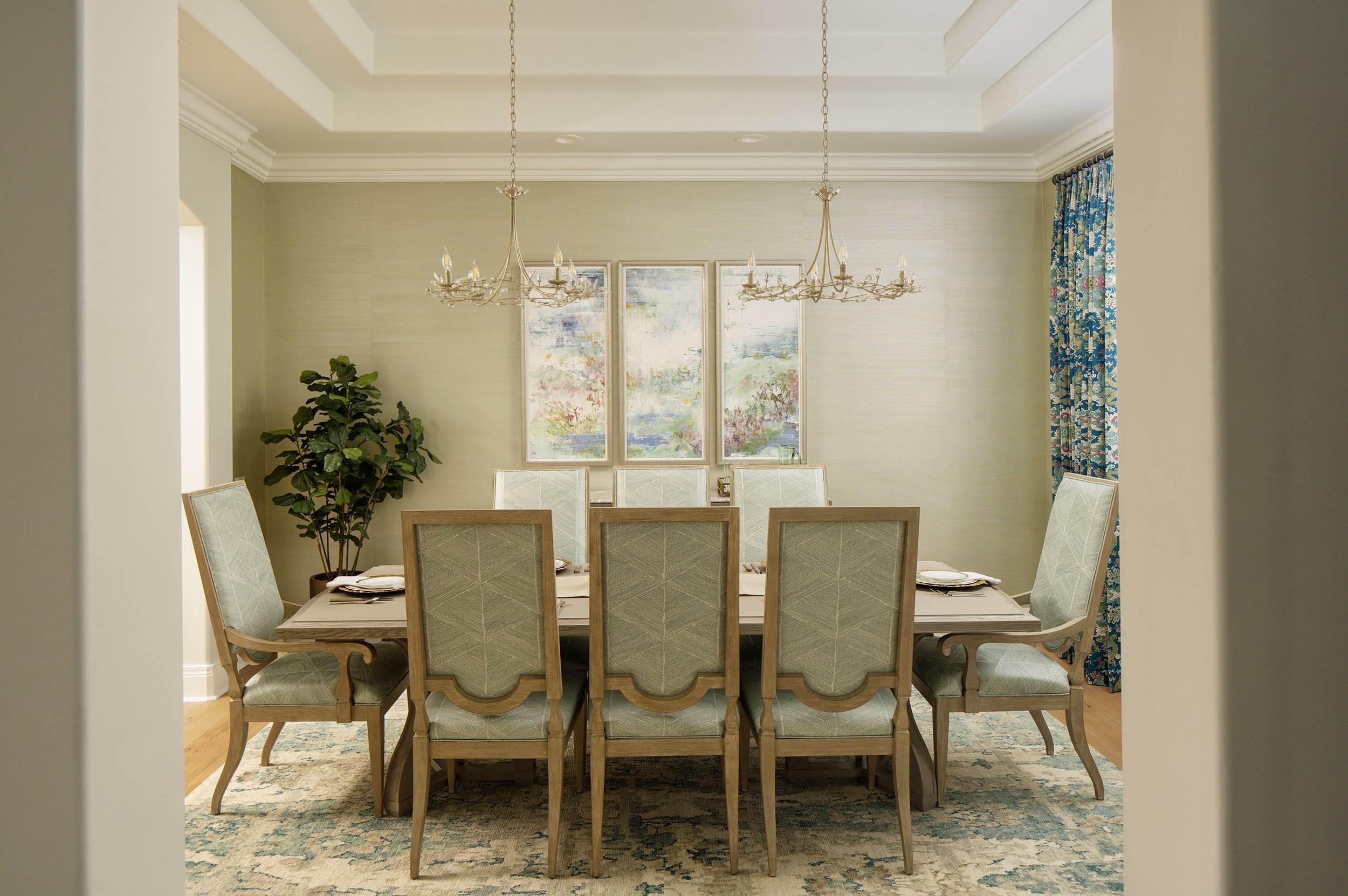 A dining room with an oval table set with plates, cups, and utensils. Eight upholstered chairs surround the table. A large potted plant is in the corner, with three framed landscape paintings on the wall behind. Two chandeliers hang from the ceiling,