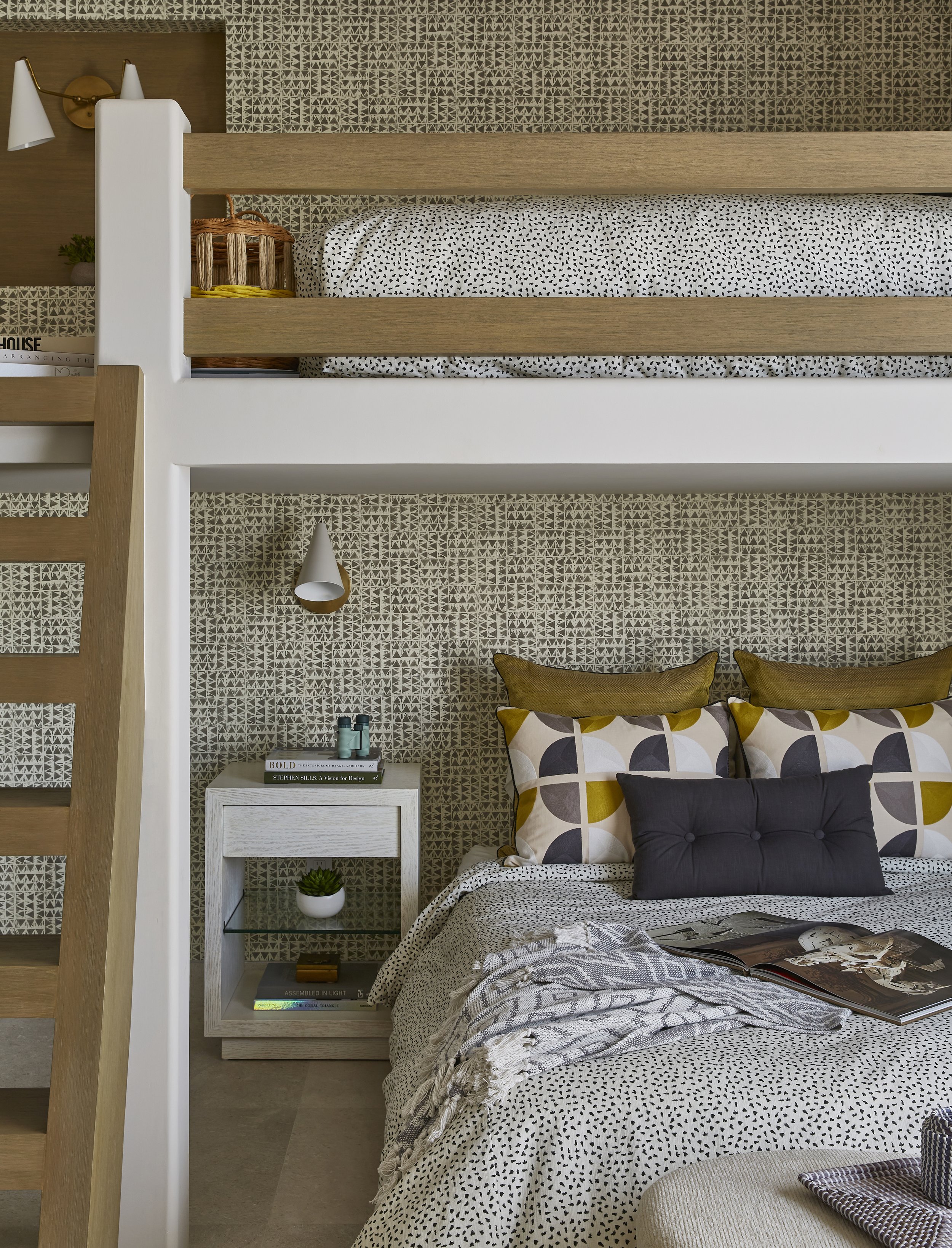 Modern bedroom with a bunk bed, patterned wallpaper, bedside table with plants and books, bed with patterned pillows, and a blanket with a magazine on top.