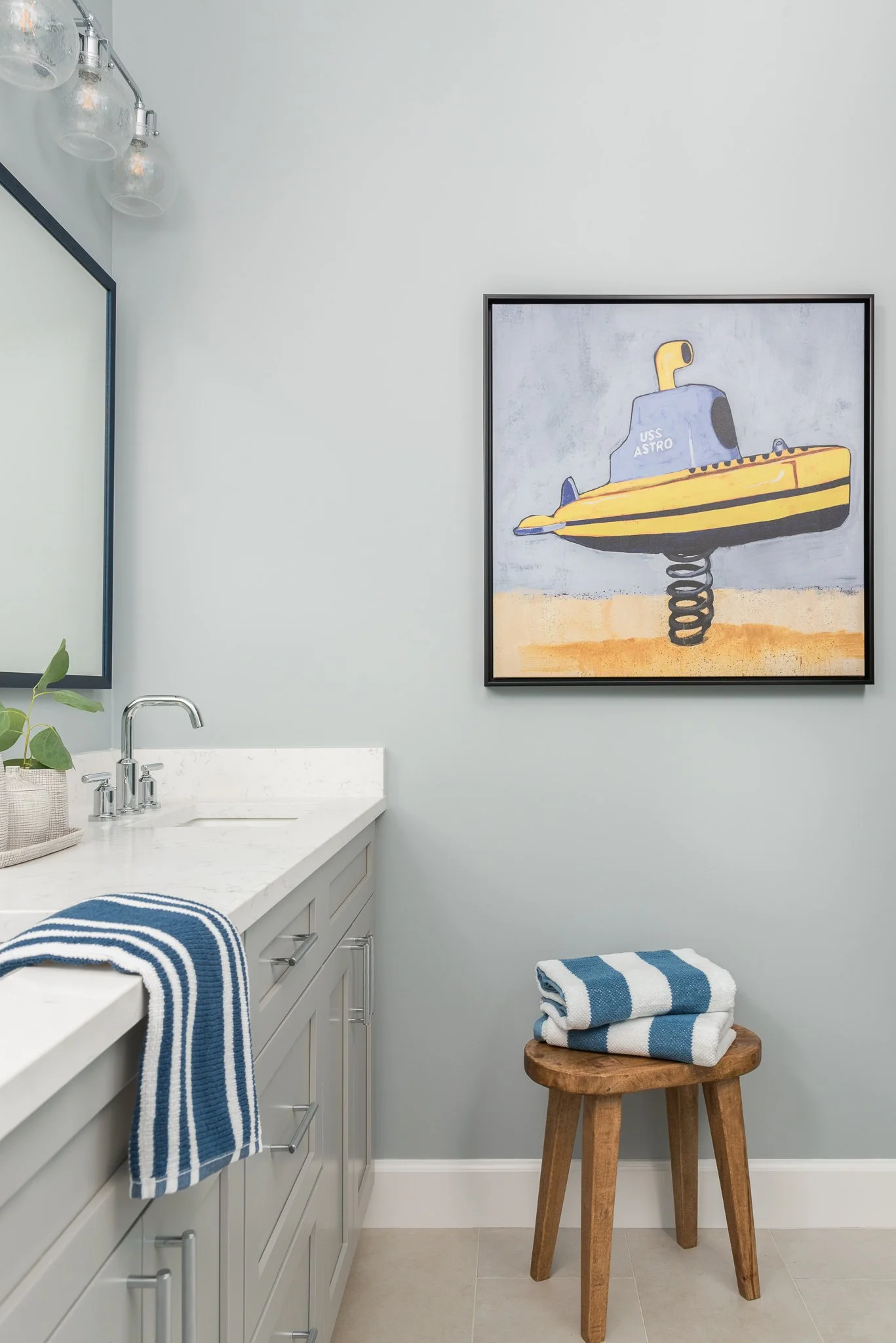 Bathroom with light gray walls, white countertop, and gray cabinetry. A mirror with black frame, a plant in a basket, and hanging glass light fixtures are visible. A blue and white striped towel hangs on the counter. A wooden stool with folded blue a