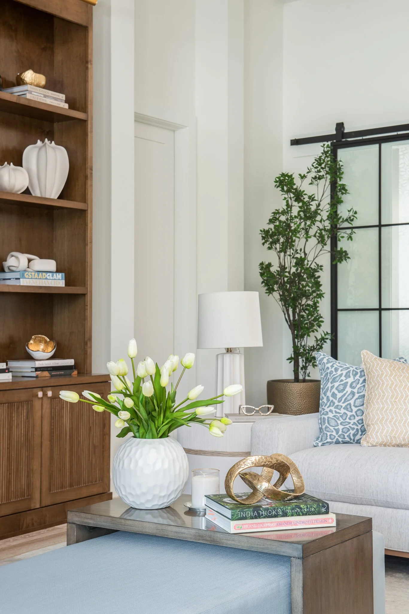 Living room with white sofa, beige and blue patterned pillows, white tulips in a white textured vase on a gray coffee table, books, gold decorative sculpture, white lamp, potted plant near a large window with black frame.